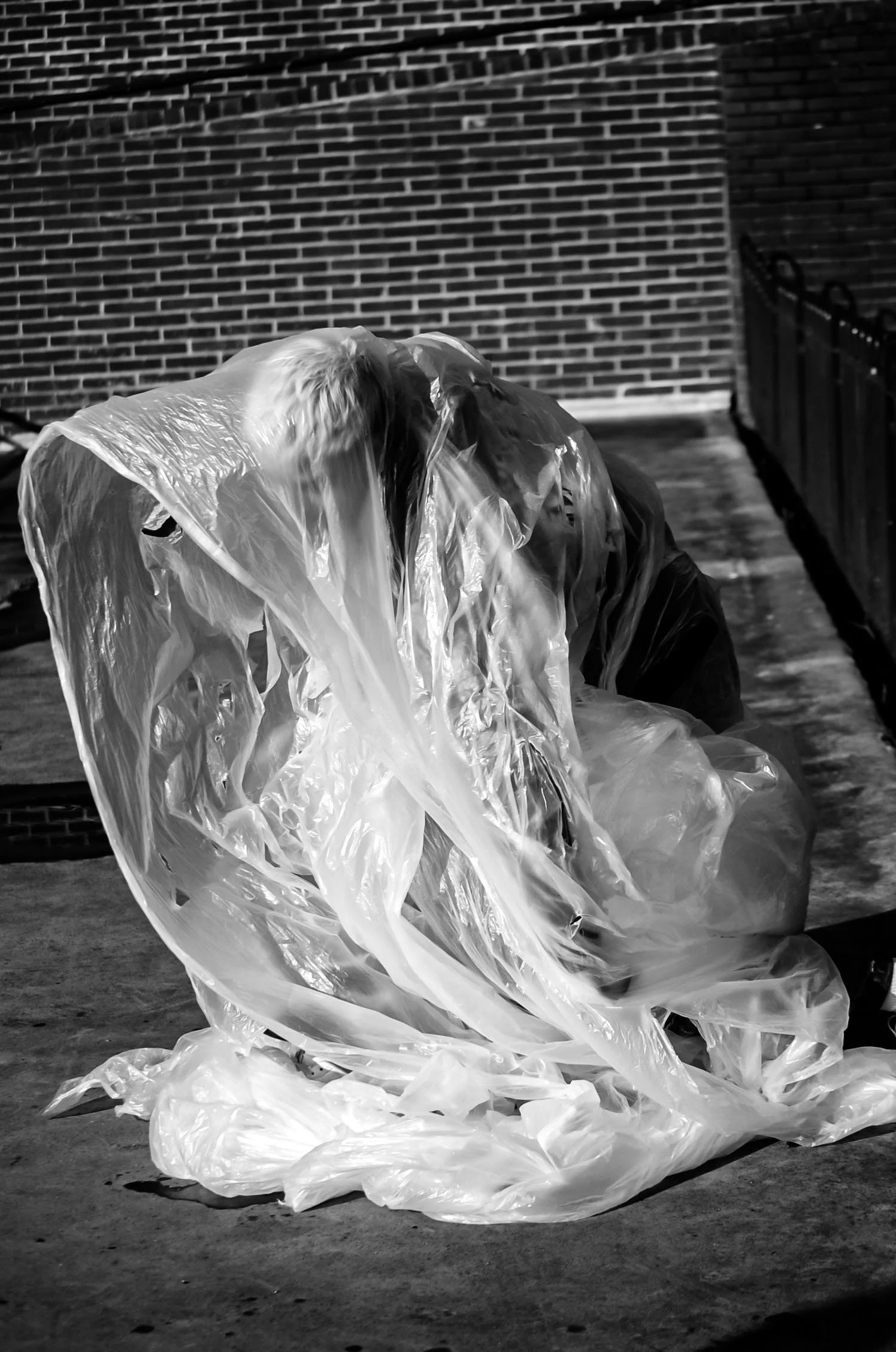 A person wearing a transparent plastic raincoat, crouched down, with a large plastic bag on the ground. The scene is outdoors, with a brick wall in the background.