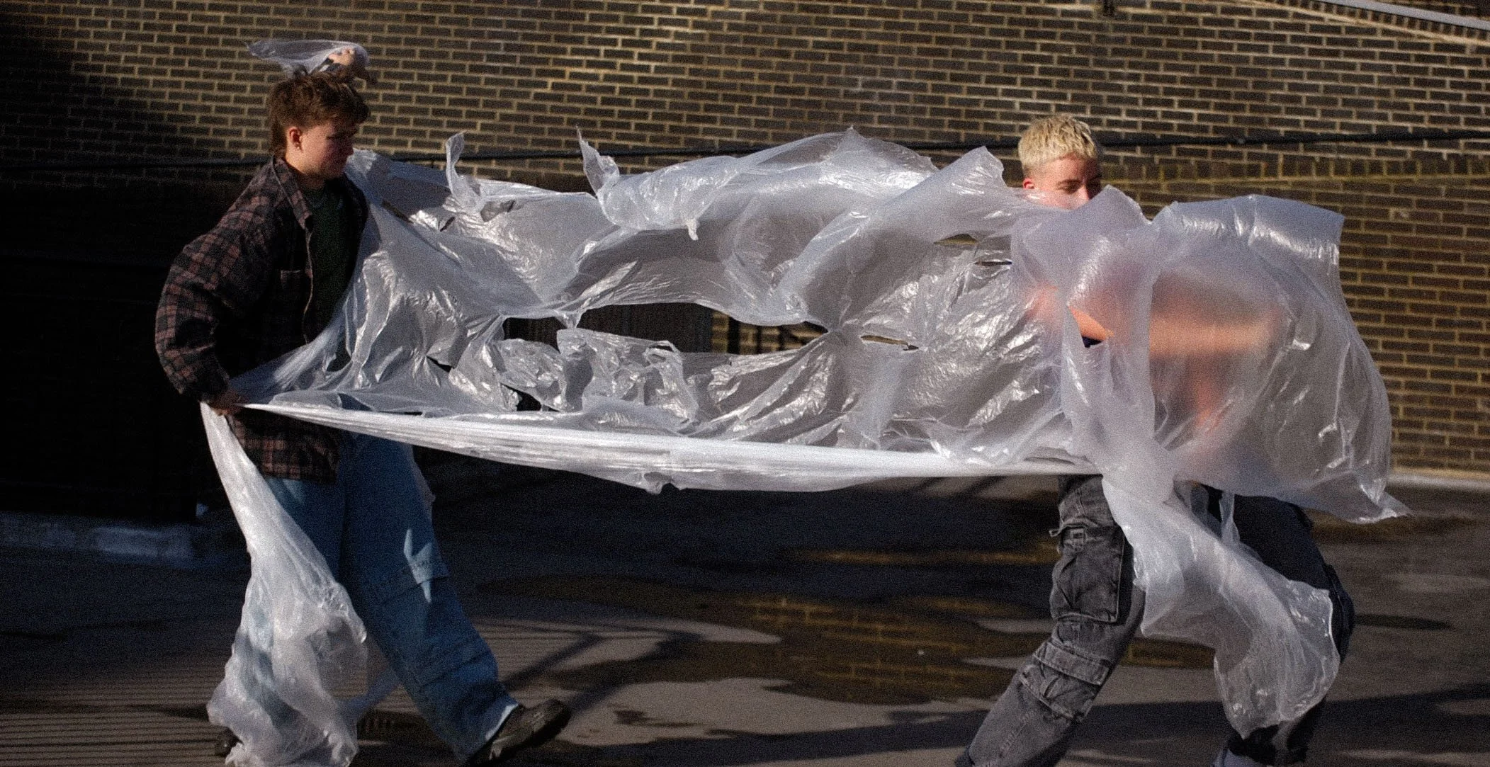 Two boys walking outside, holding a large clear plastic sheet between them.