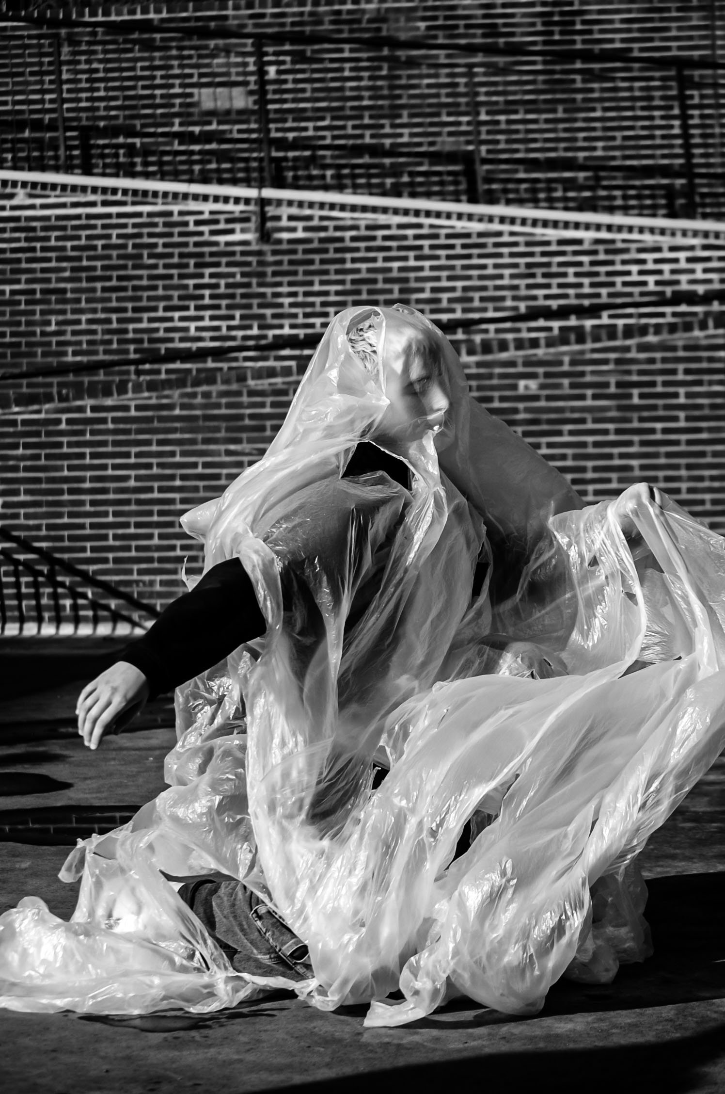 A person wearing a plastic raincoat is seated on the ground, surrounded by large plastic bags, against a background of a brick wall and an outdoor staircase.