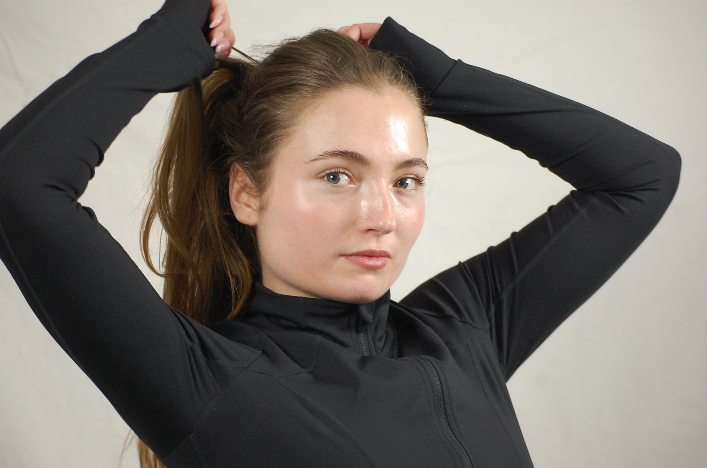 Young woman with long brown hair, blue eyes, and fair skin, dressed in a black long-sleeve athletic shirt, tying her hair back against a plain light background.