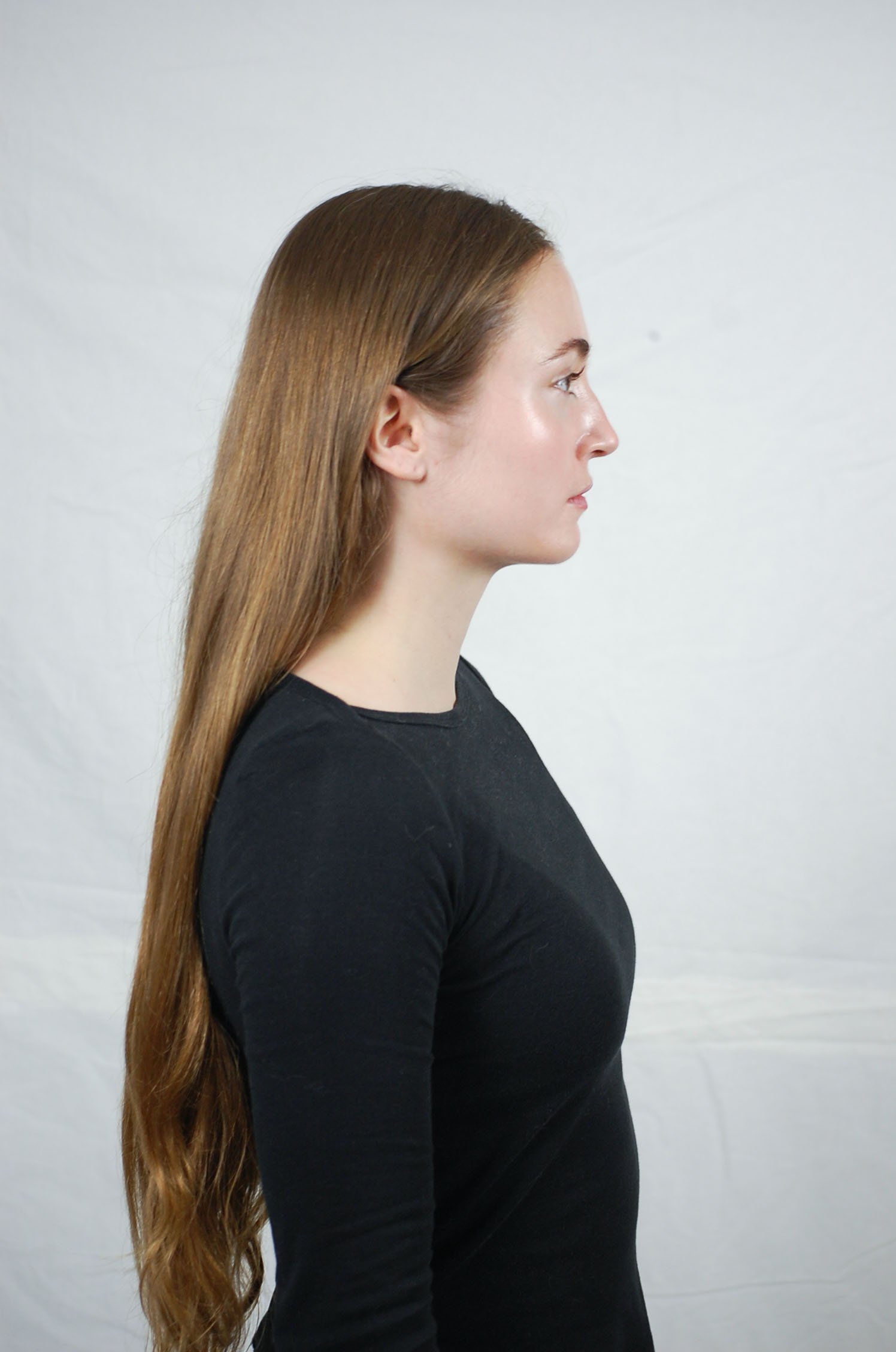 Side profile of a woman with long, straight, brown hair wearing a black top, standing against a plain light-colored background.