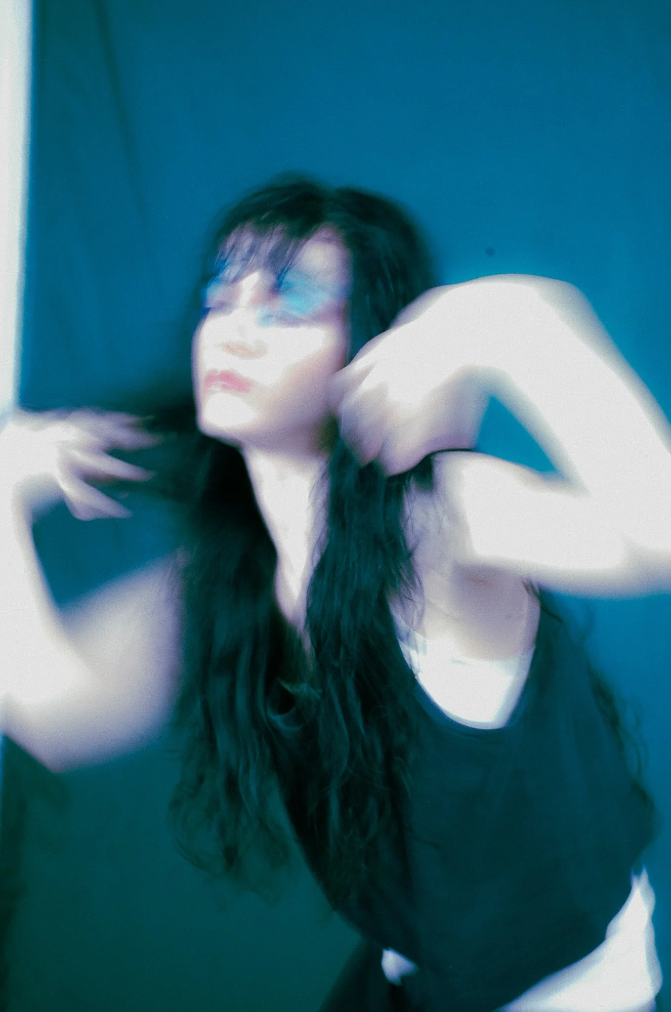 A woman with long dark hair, wearing glasses, posing with her hand behind her head against a blue background.