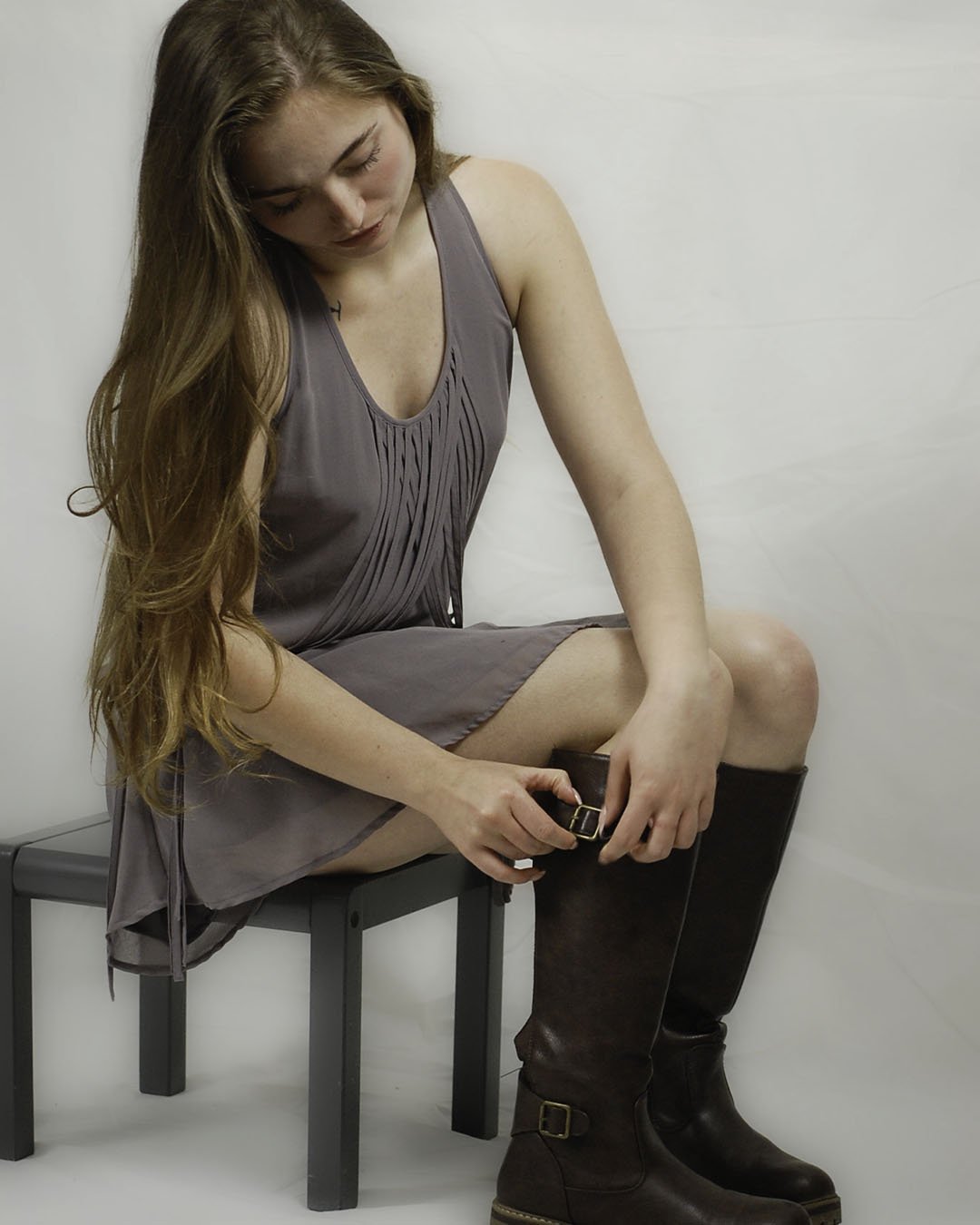 Young woman sitting on a gray bench, adjusting her brown leather riding boots, wearing a gray dress, with long red hair, against a plain white wall.