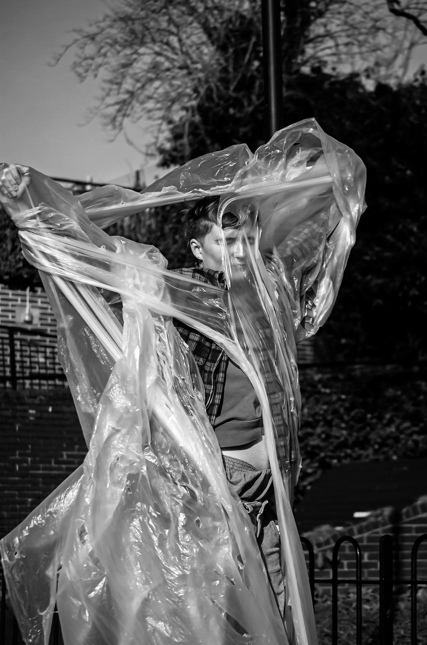 A person wearing a plaid jacket and striped pants holding a large, transparent plastic sheet in an outdoor setting, with trees and a brick wall in the background.