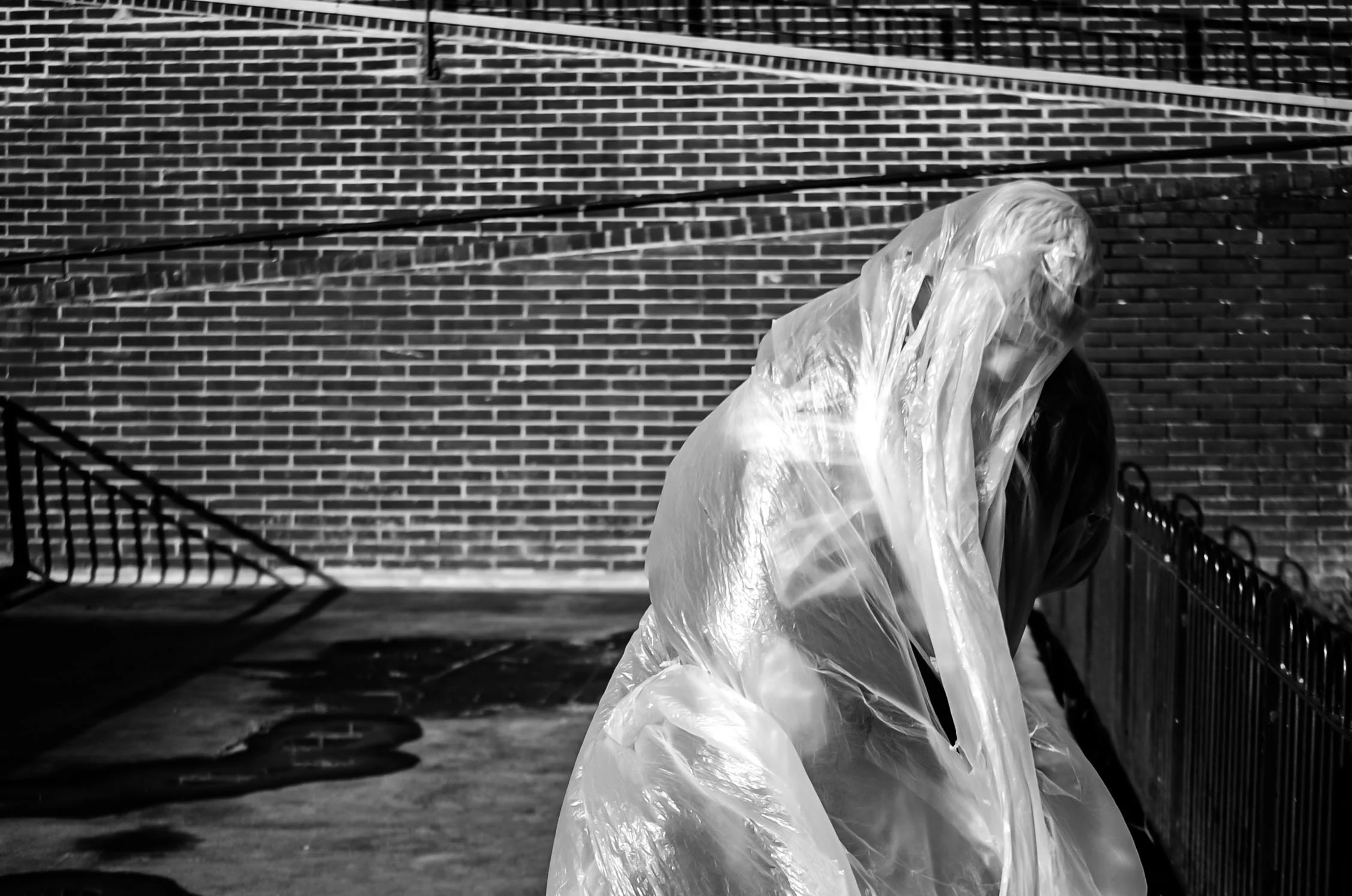 A person wearing a translucent plastic raincoat standing outdoors near brick walls and metal railings in black and white.