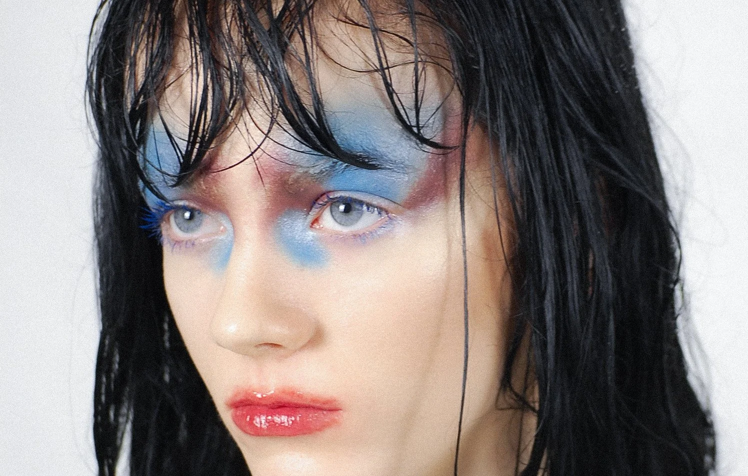 Close-up of a person with wet black hair and light makeup, wearing vibrant blue eyeshadow and shiny pink lipstick.