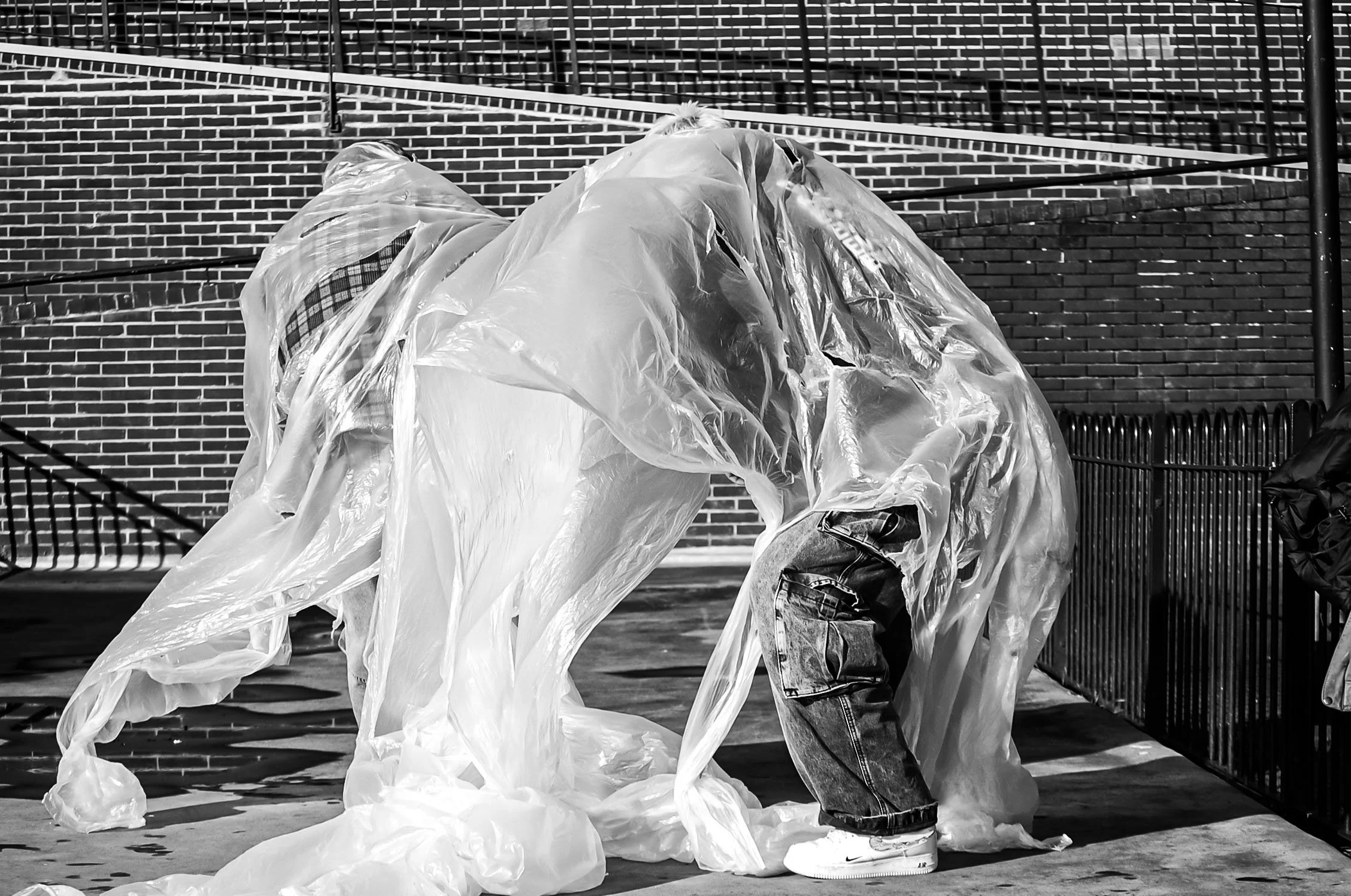 A person wearing jeans and a jacket is standing outdoors, handling plastic sheeting against a brick wall with a fence in the background.