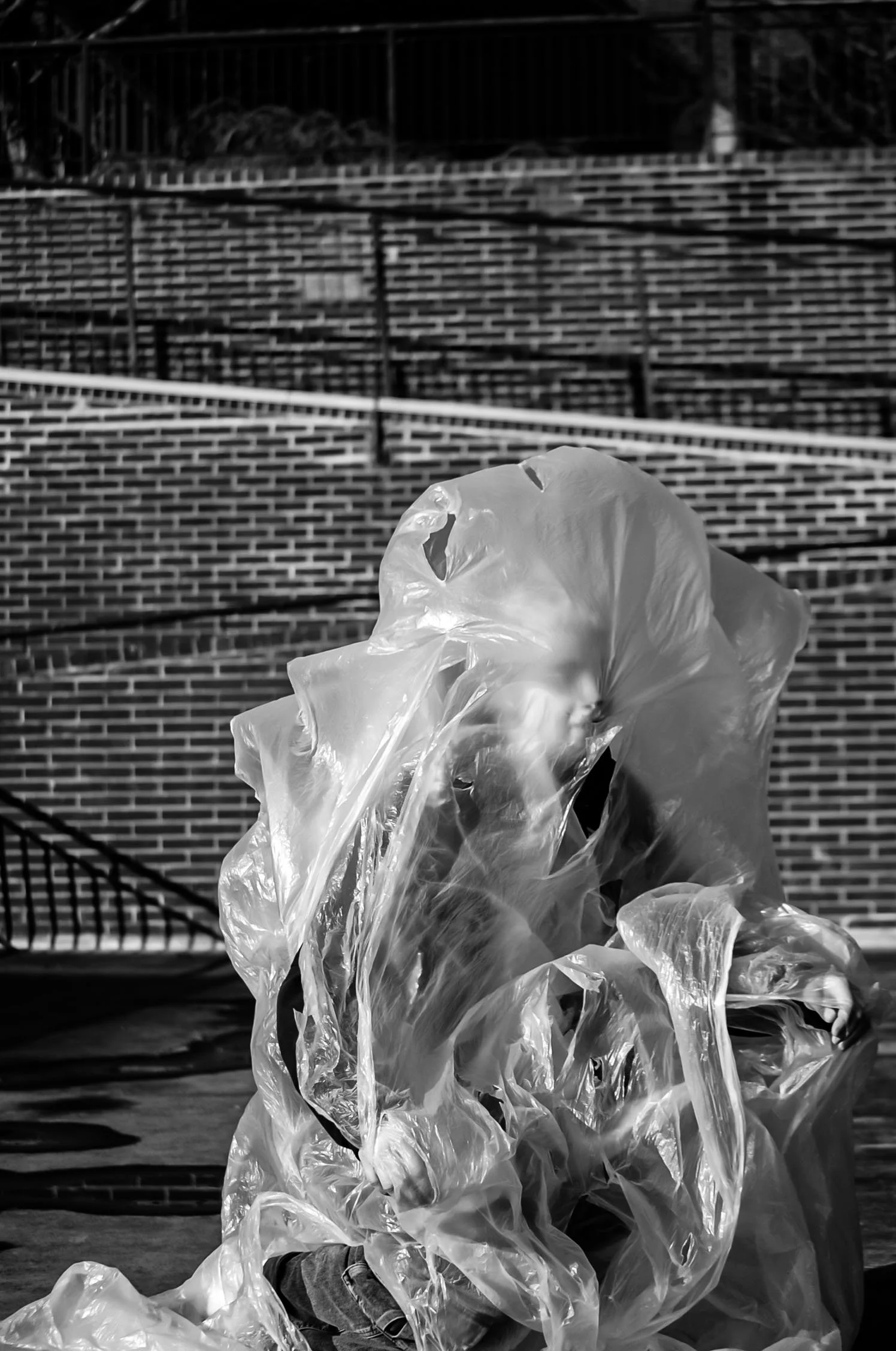 Close-up of a person sitting on the ground, wrapped in a transparent plastic bag, with a brick wall and metal stairs in the background.