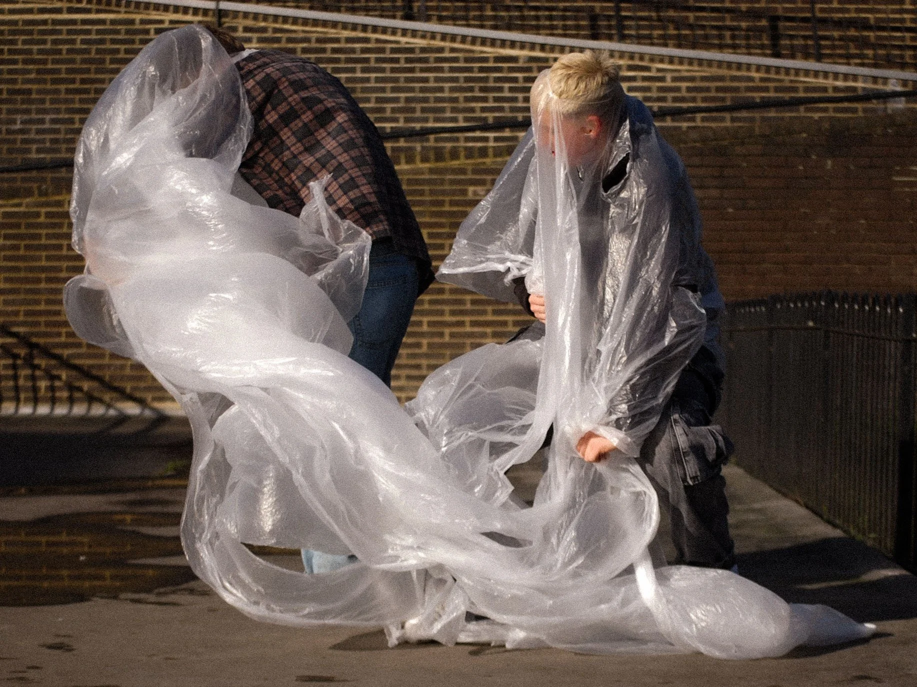 Two people working with large clear plastic sheets outdoors at night, with a brick wall and a fence in the background.
