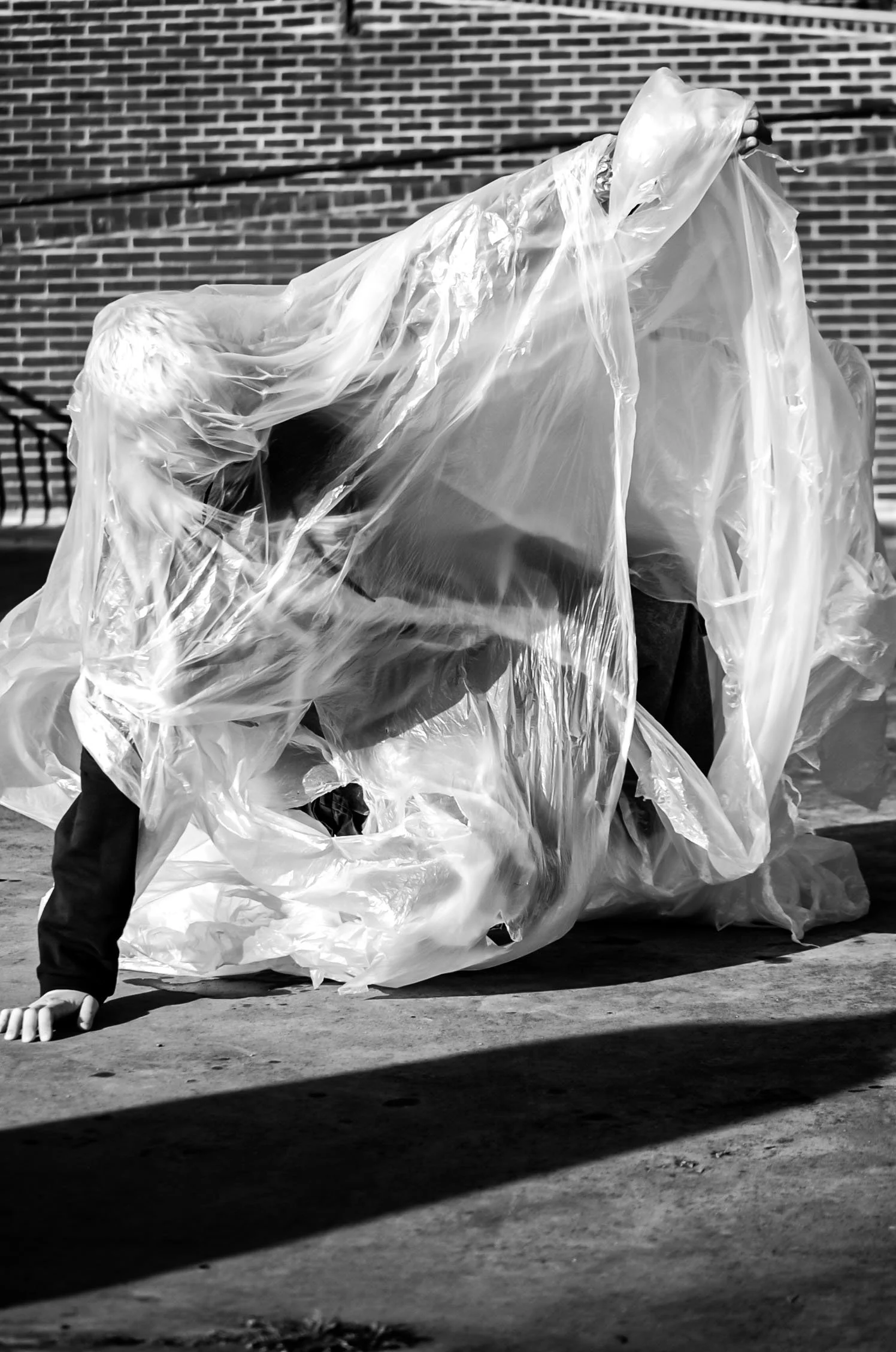 Person crouching behind large plastic trash bags, with a brick wall in the background, in black and white.