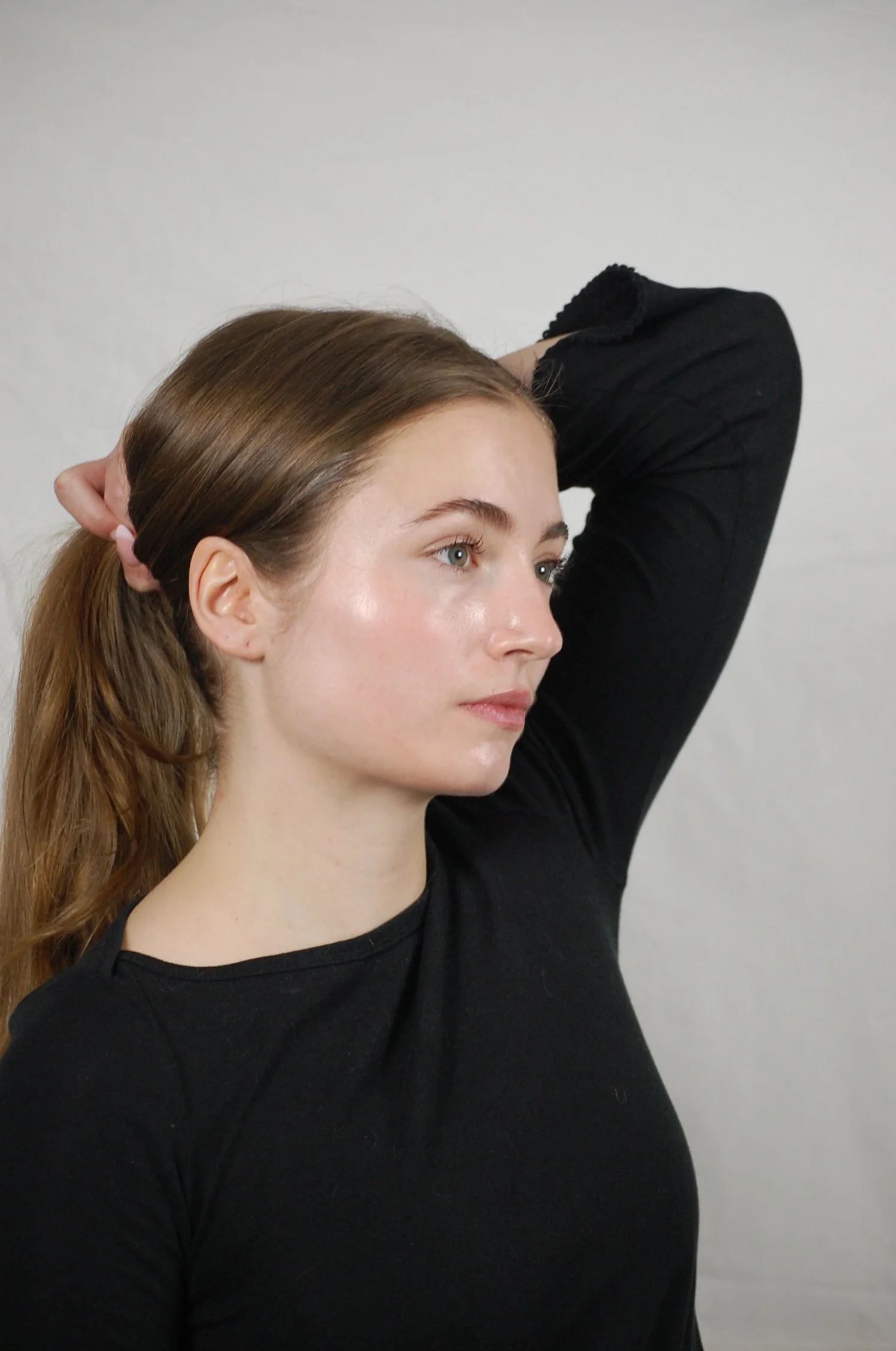 A woman with long brown hair tied back, wearing a black shirt, poses with her right arm bent and her hand resting behind her head, standing against a plain light-colored background.