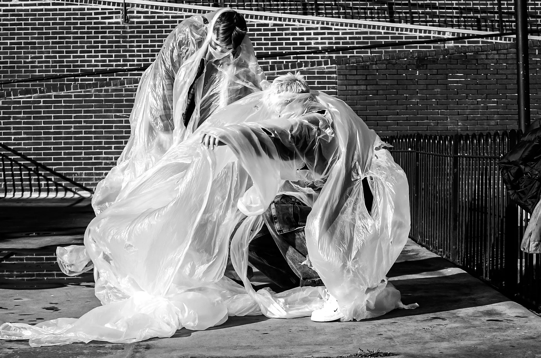 Two people are kneeling on the sidewalk, wearing plastic rain ponchos, with a brick wall and metal railing in the background. They appear to be wrapping or unwrapping something with plastic.