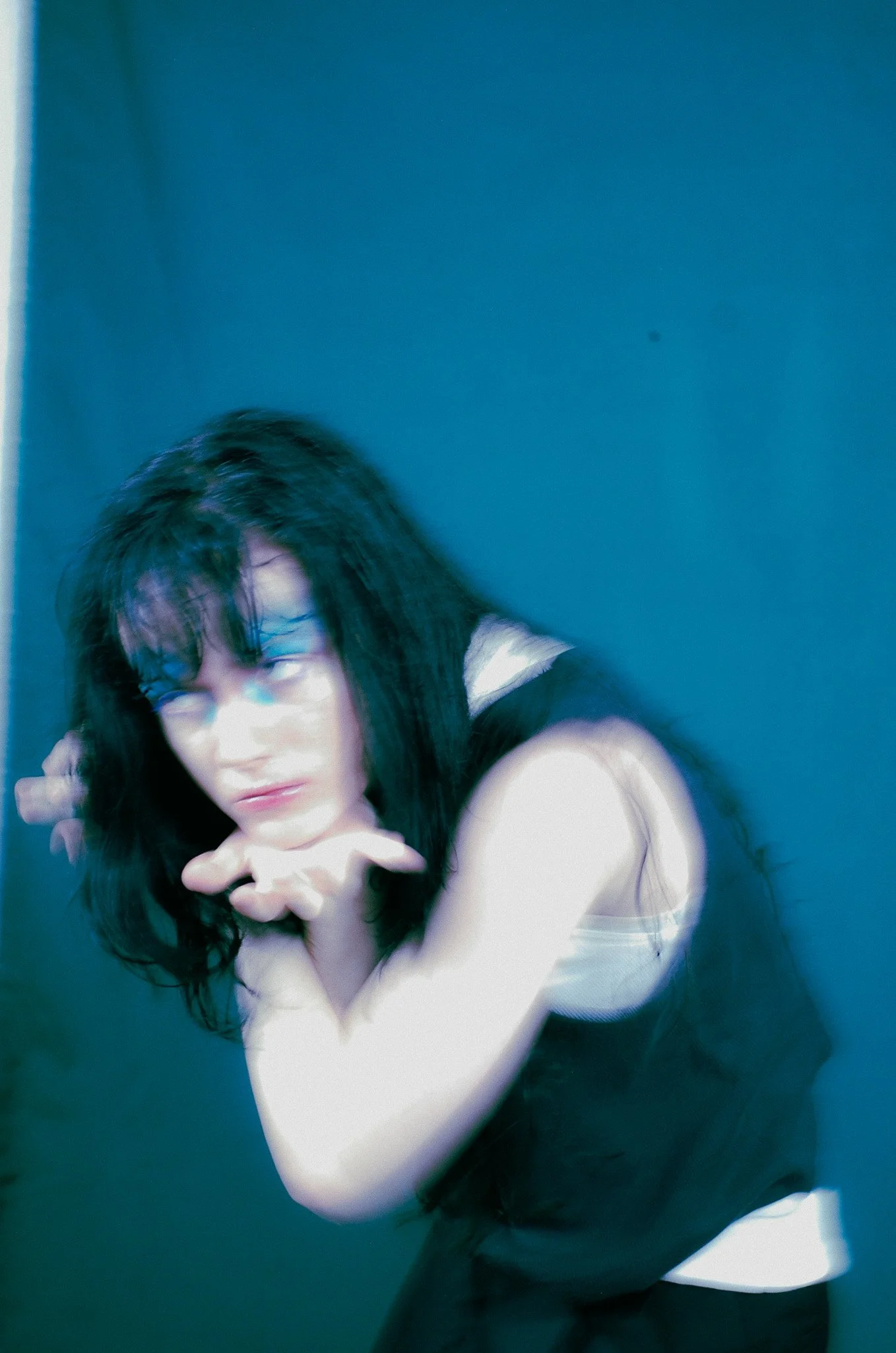 A woman with long dark hair and glasses in a white top, posing against a blue background with her hand near her face.