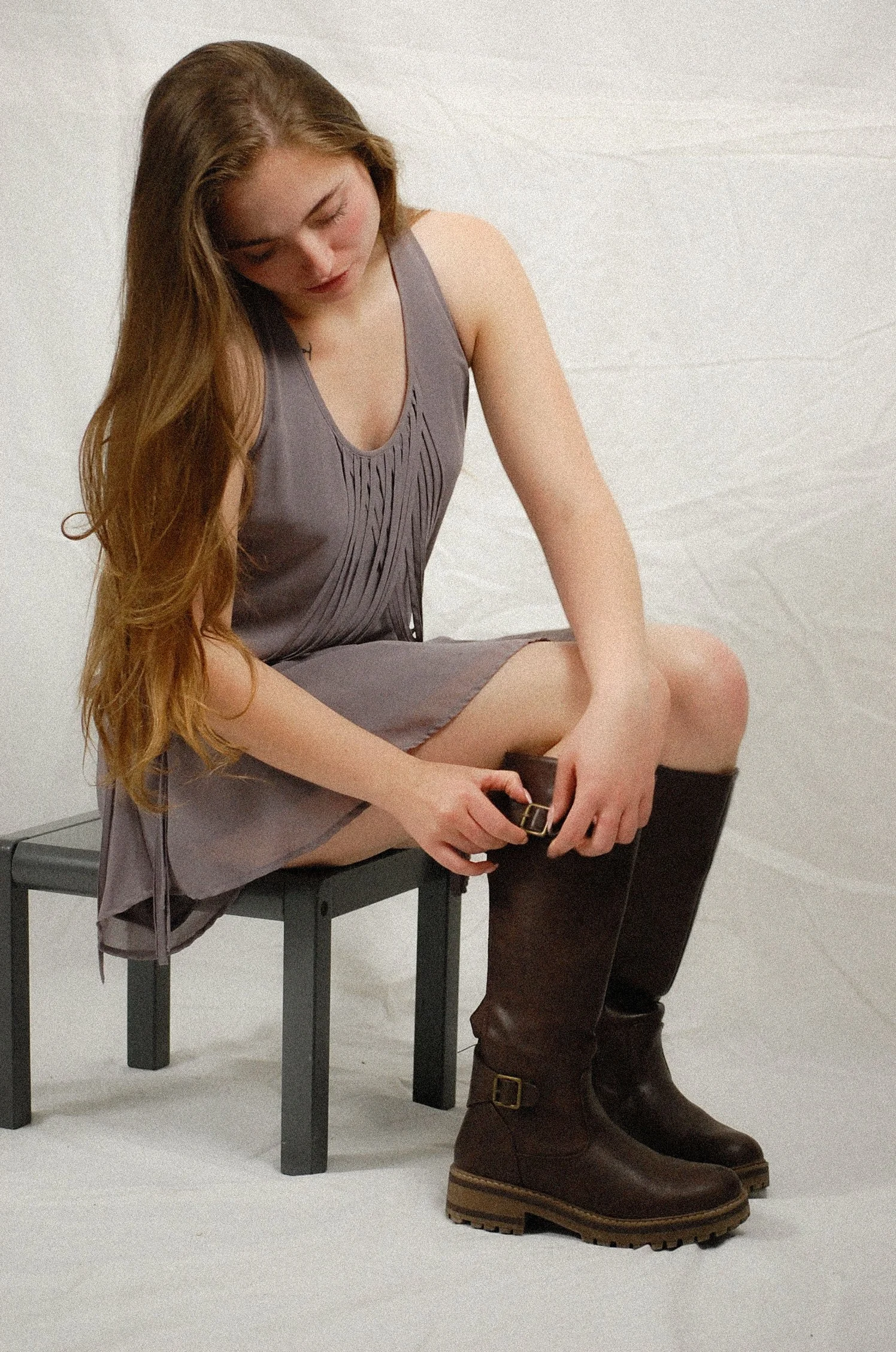 A young woman with long, wavy, light brown hair sitting on a black stool in front of a plain white background. She wears a sleeveless, gray dress with pleated detail in the front, black knee-high boots with chunky heels and buckles. She is looking down, with her head slightly tilted, and appears to be adjusting her boot.