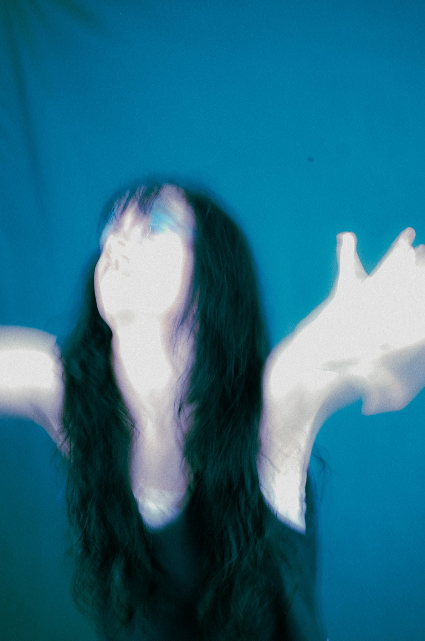 A person with long dark hair raising their arms, with a blue background and light effects creating a surreal or abstract appearance.