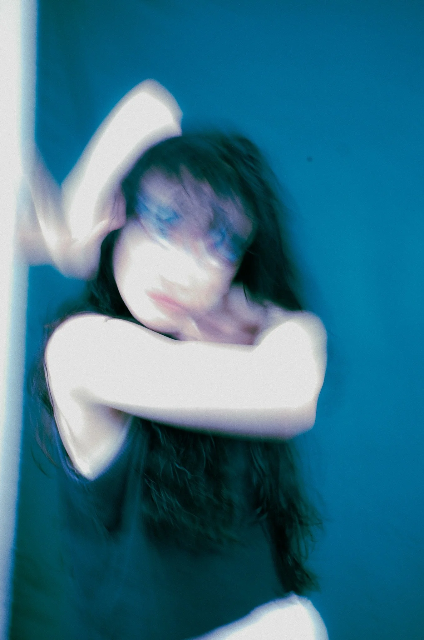Blurred portrait of a woman with dark hair, standing against a blue wall, with her eyes closed and hand touching her head.