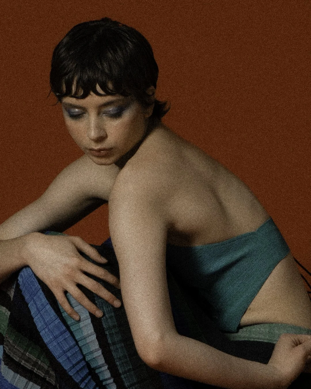 A young woman with short dark hair and blue eyeshadow, sitting against an orange background, wearing a strapless turquoise top and patterned pants, looking downward with her eyes closed.