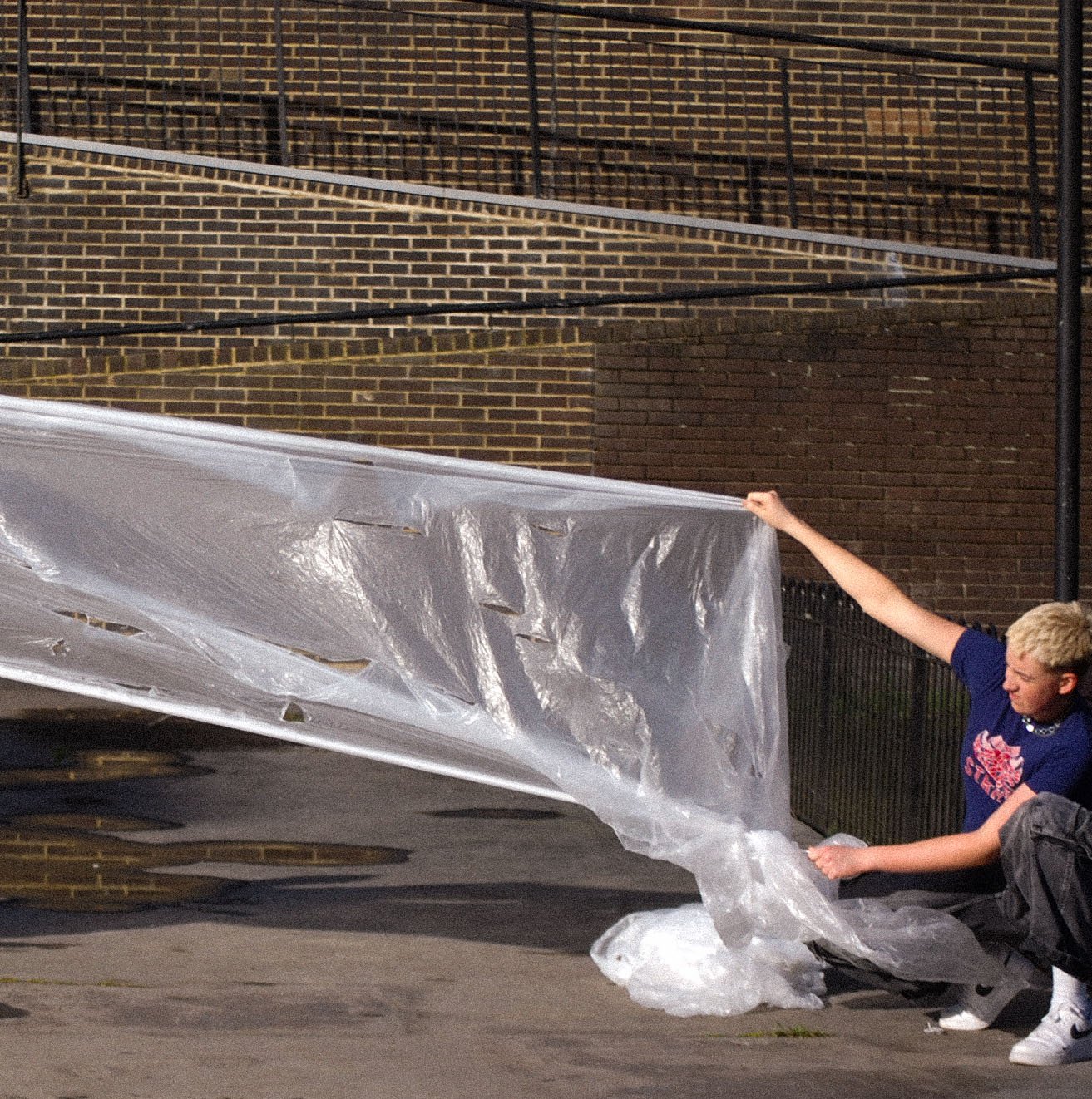 Two children are holding a large, clear plastic sheet against a brick wall. They are outside near a sidewalk, with a black fence and stairs visible in the background.