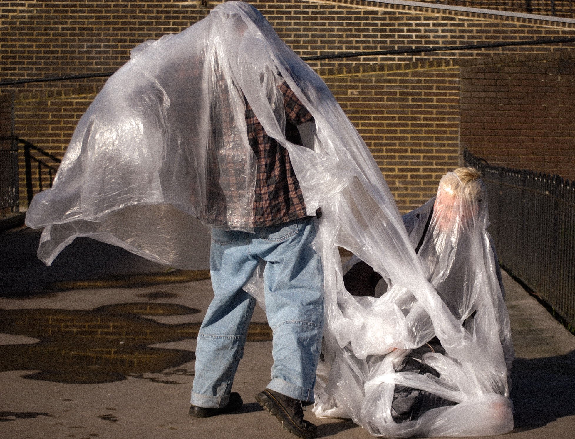 Person wearing a plaid shirt and jeans, standing on a sidewalk, covered with a large clear plastic bag with more bags on the ground, against a brick wall background.