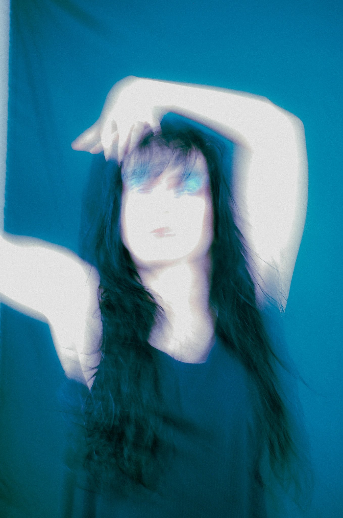 Person with long dark hair, shirtless, against a blue background, with hands behind head.