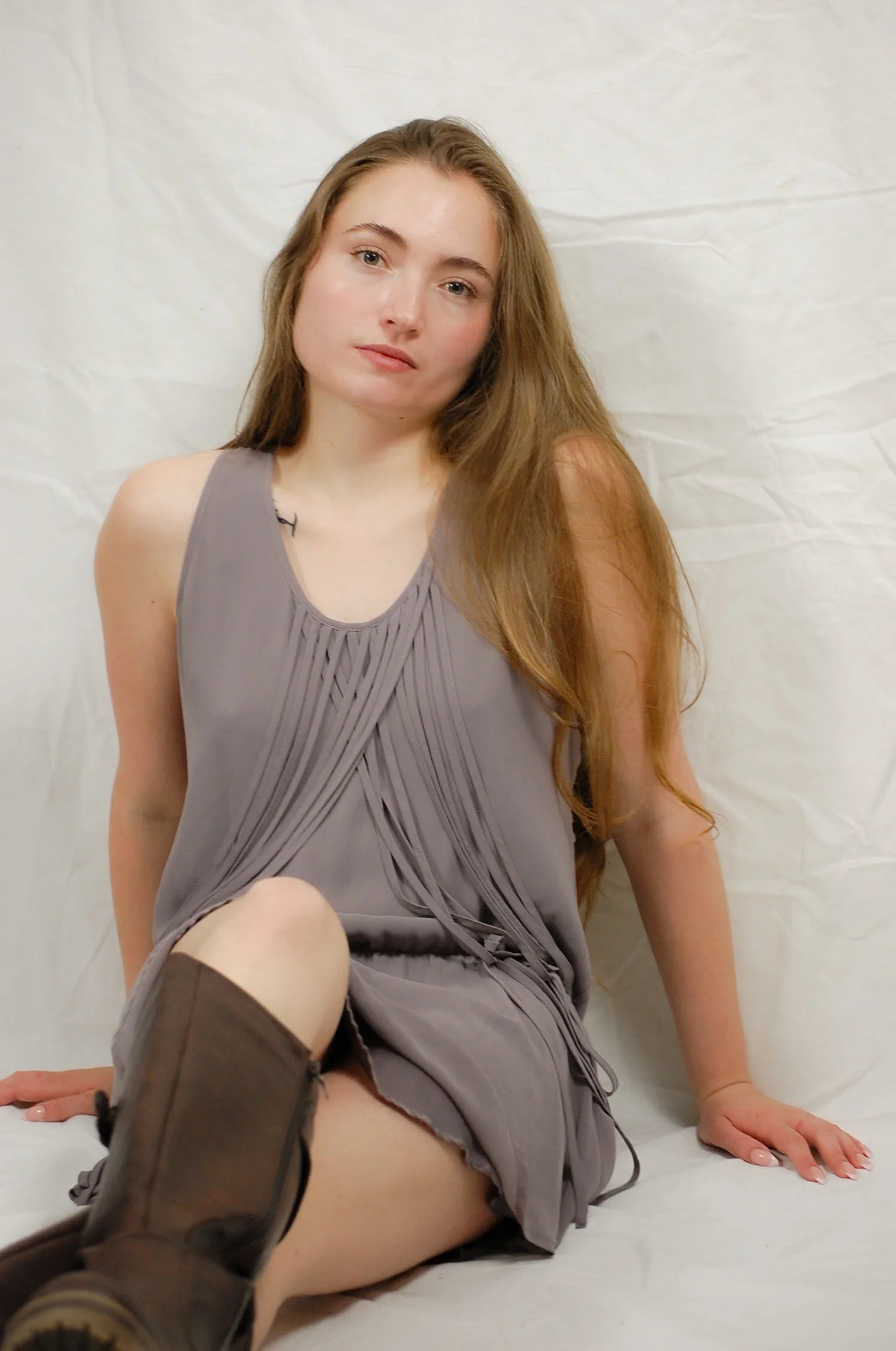 A young woman with long, light brown hair and fair skin sitting on a white surface against a white background. She is wearing a sleeveless, gray top and matching skirt with pleats, and brown knee-high boots. She gazes at the camera with a neutral exp