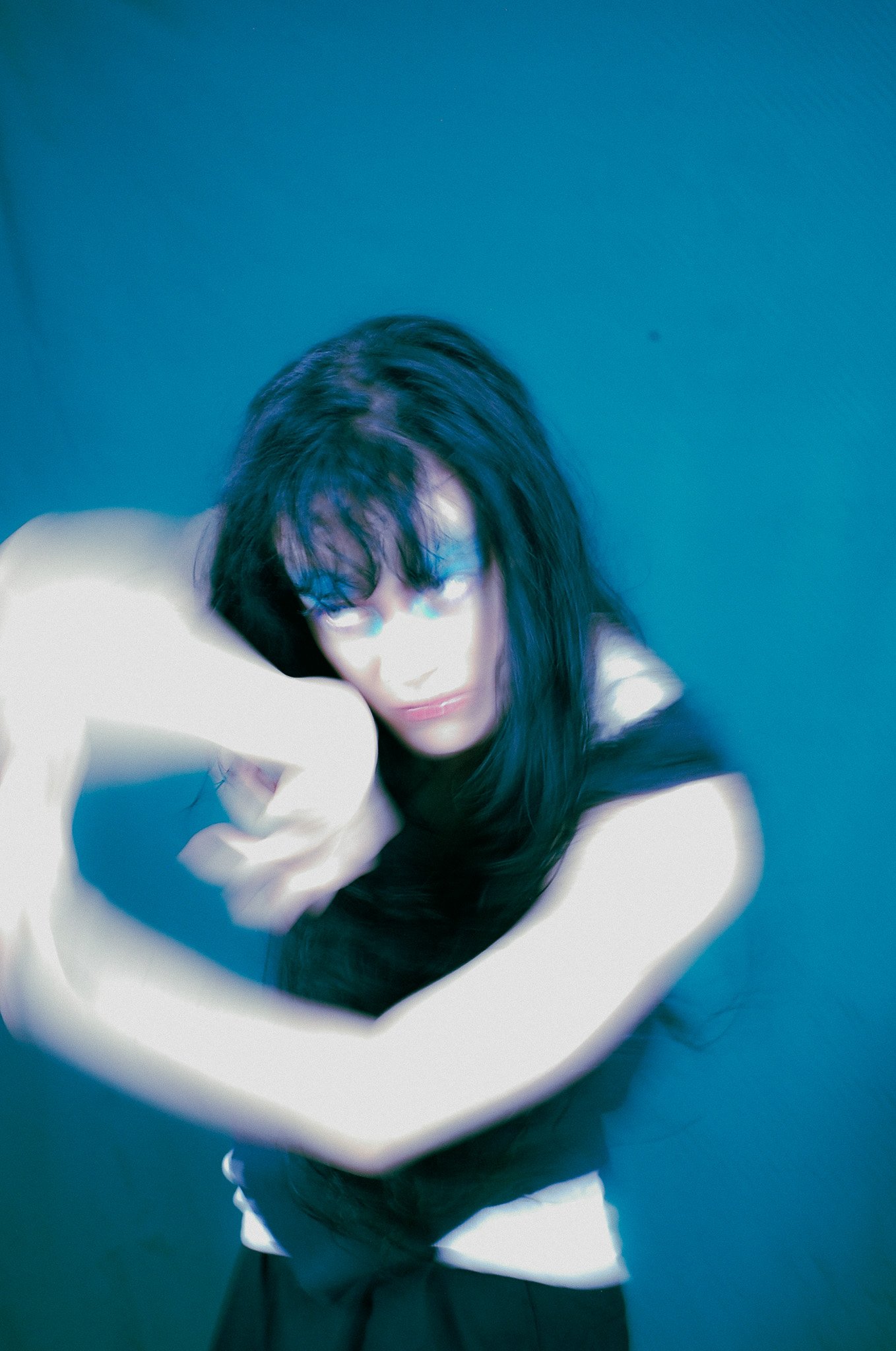 A woman with dark hair and glasses takes a selfie against a blue wall, wearing a white top.
