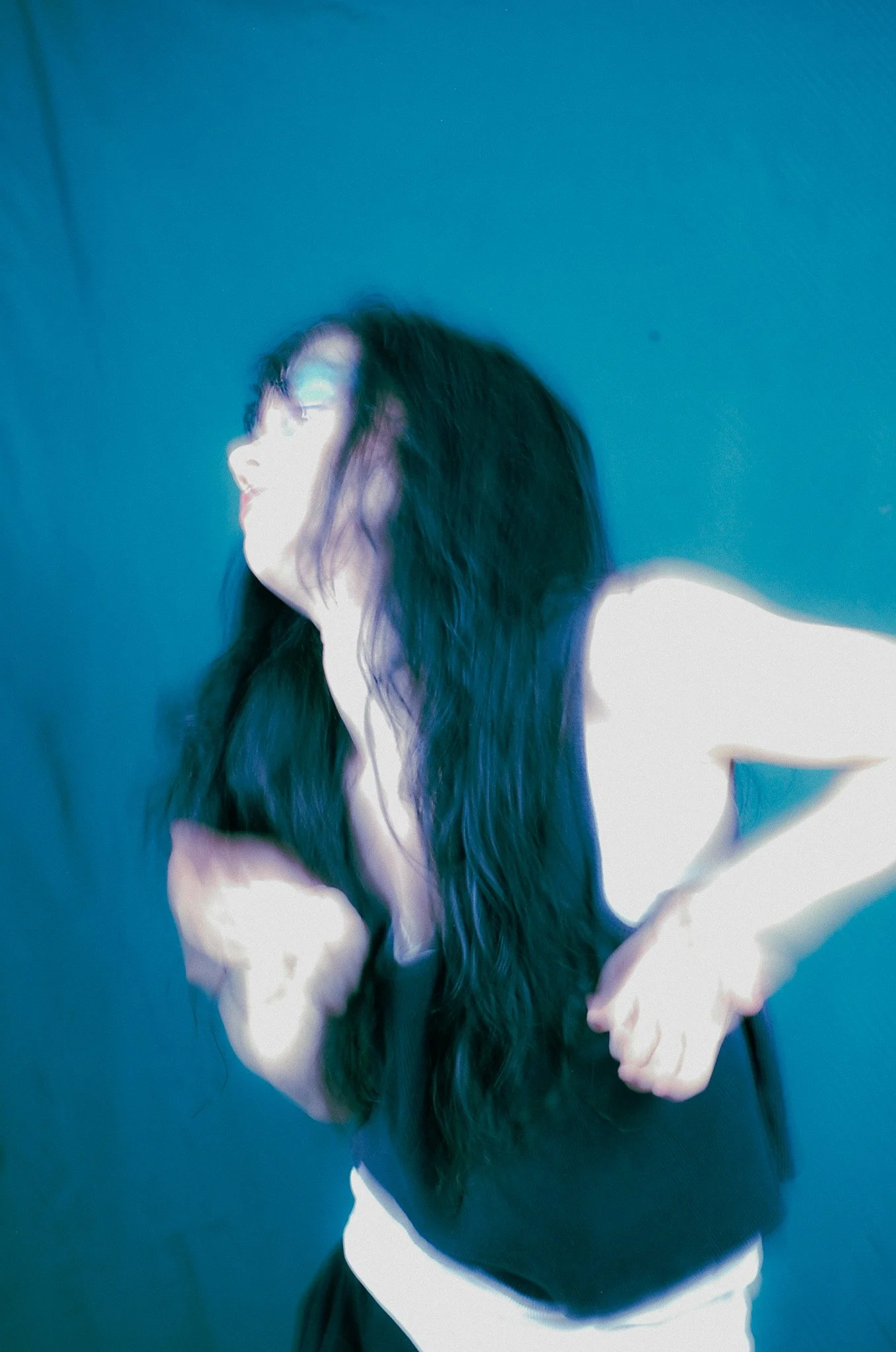 A woman with long, dark hair is standing in front of a dark teal wall, smiling and looking to her left. She is wearing a sleeveless white dress.