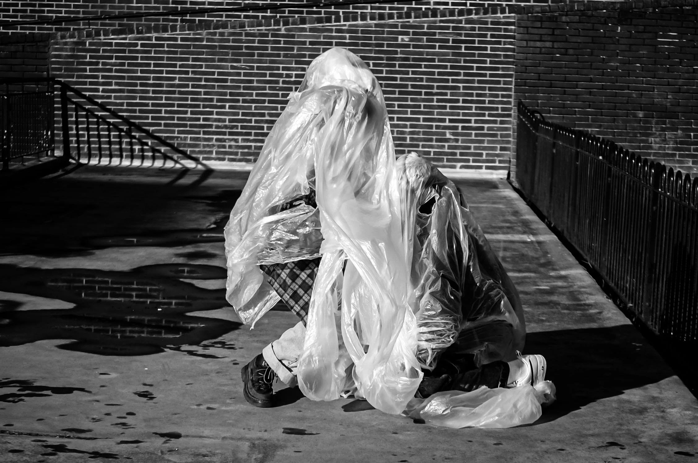 A person kneeling on the ground, wearing oversized black shoes, surrounded by crumpled plastic bags, in front of a brick wall. Shadows cast by a fence are visible on the ground.