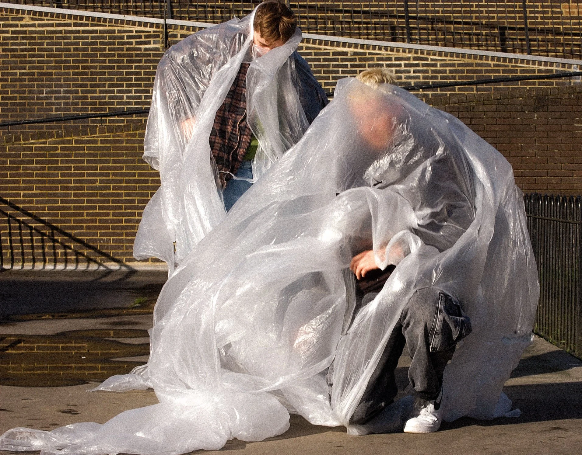 Two people kneeling on the sidewalk, handling a large transparent plastic sheet near a brick wall at night.