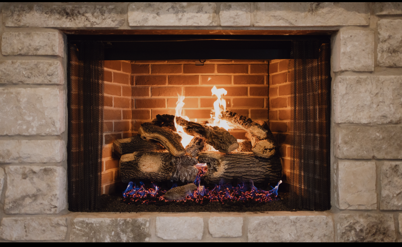 A fireplace with burning logs and visible flames, surrounded by a brick interior and a stone mantel.