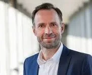 Et mandet portræt med mellemlangt, mørkt hår og skæg, iført hvid skjorte og mørk blazer, stående foran en kontorbygning med glasfacade.