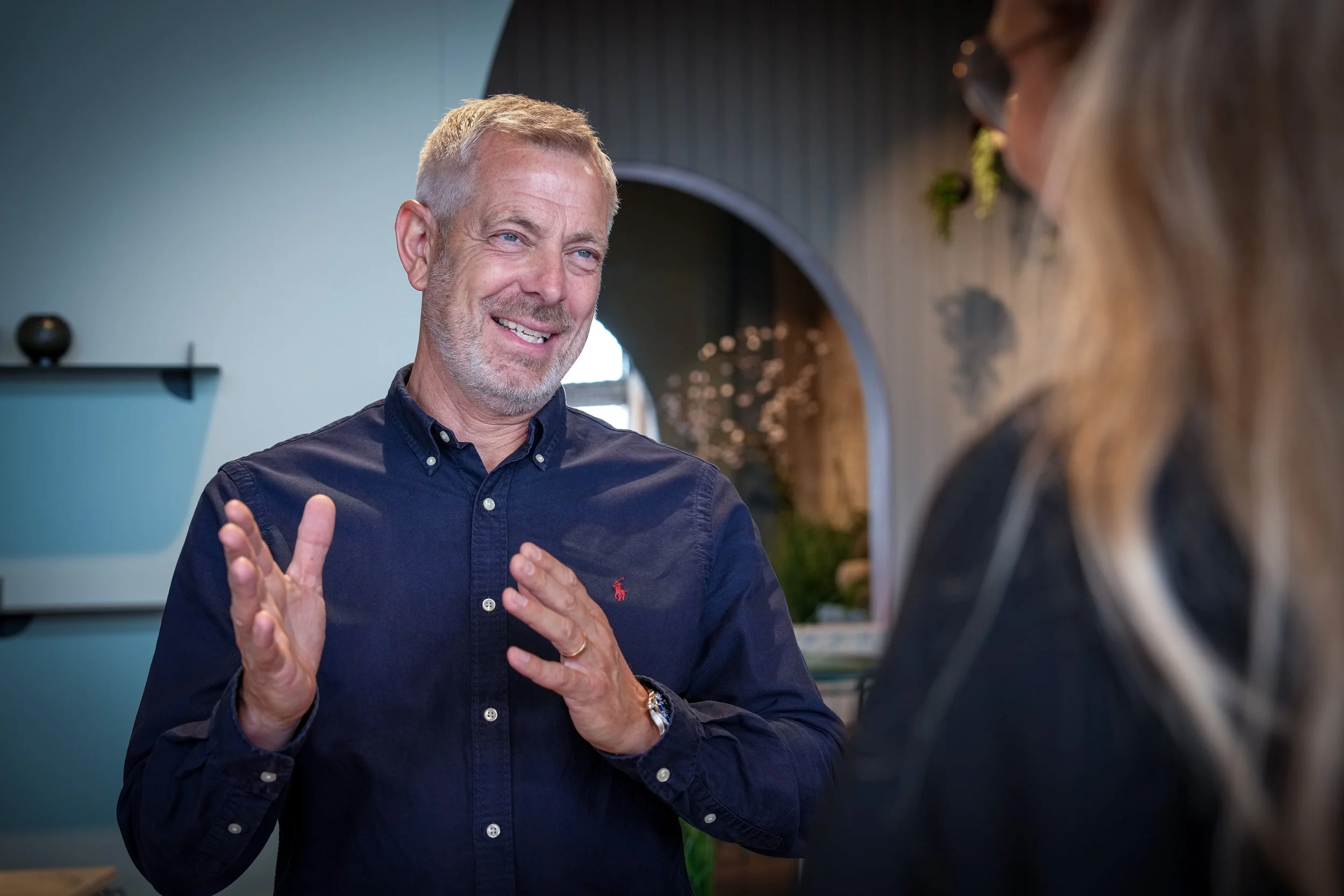 En mand med blond kort hår og gråt skæg smiler og taler med en kvinde med brunt hår og solbriller i et moderne interiør.