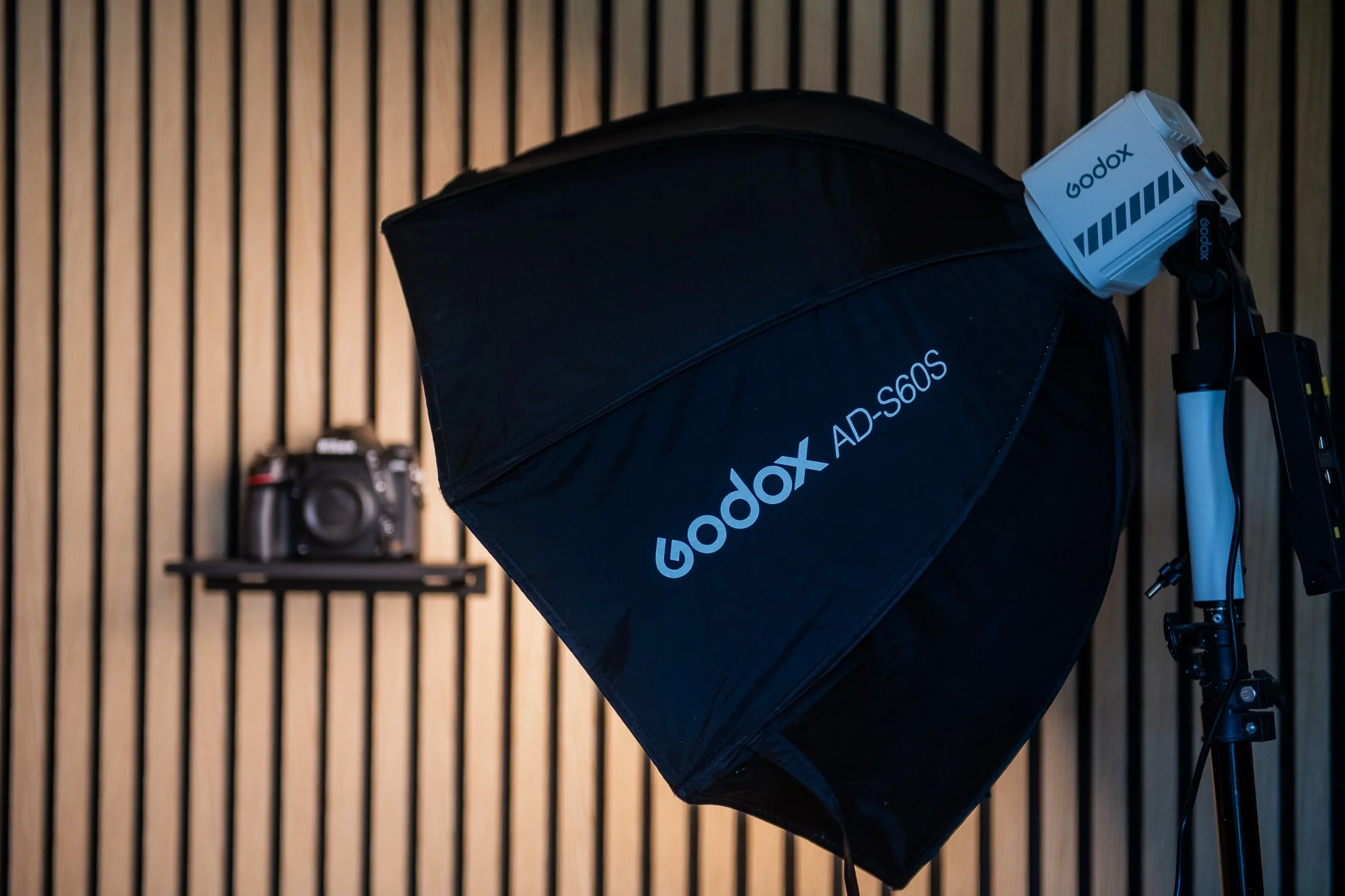 Studio photography setup with a Godox AD-S60S softbox, camera in the background, and lighting equipment against a wooden slatted wall.