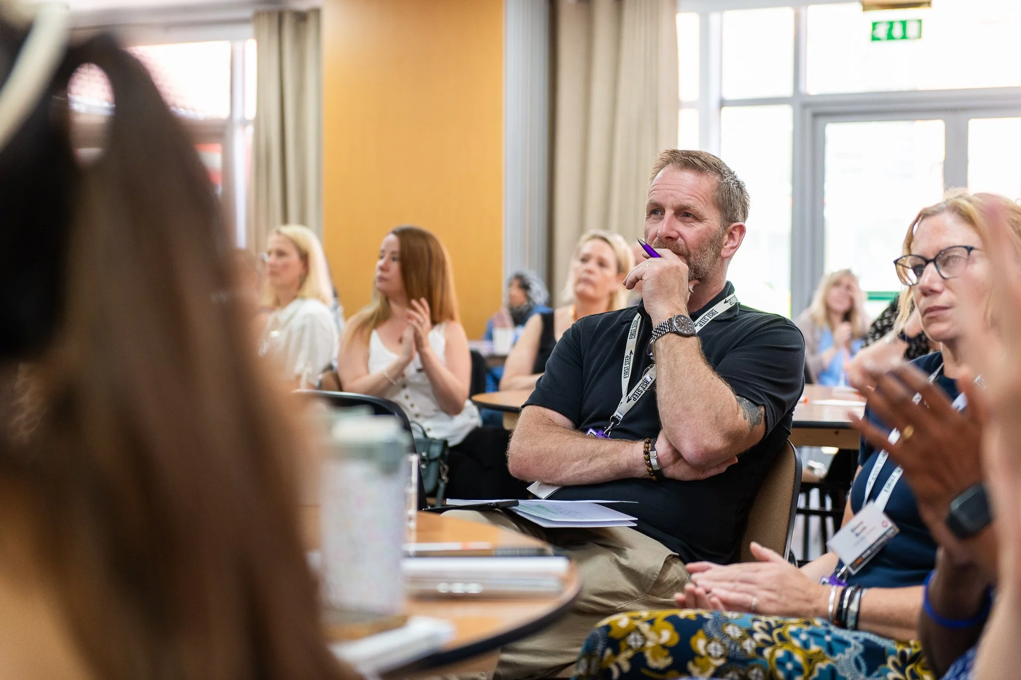 A group of people attending a conference or seminar in a well-lit room with large windows and beige walls. One man with a beard and short hair, wearing a black t-shirt and lanyard, is resting his chin on his hand, appearing attentive. Other attendees, mostly women, are seated around tables, listening to a speaker or presentation.