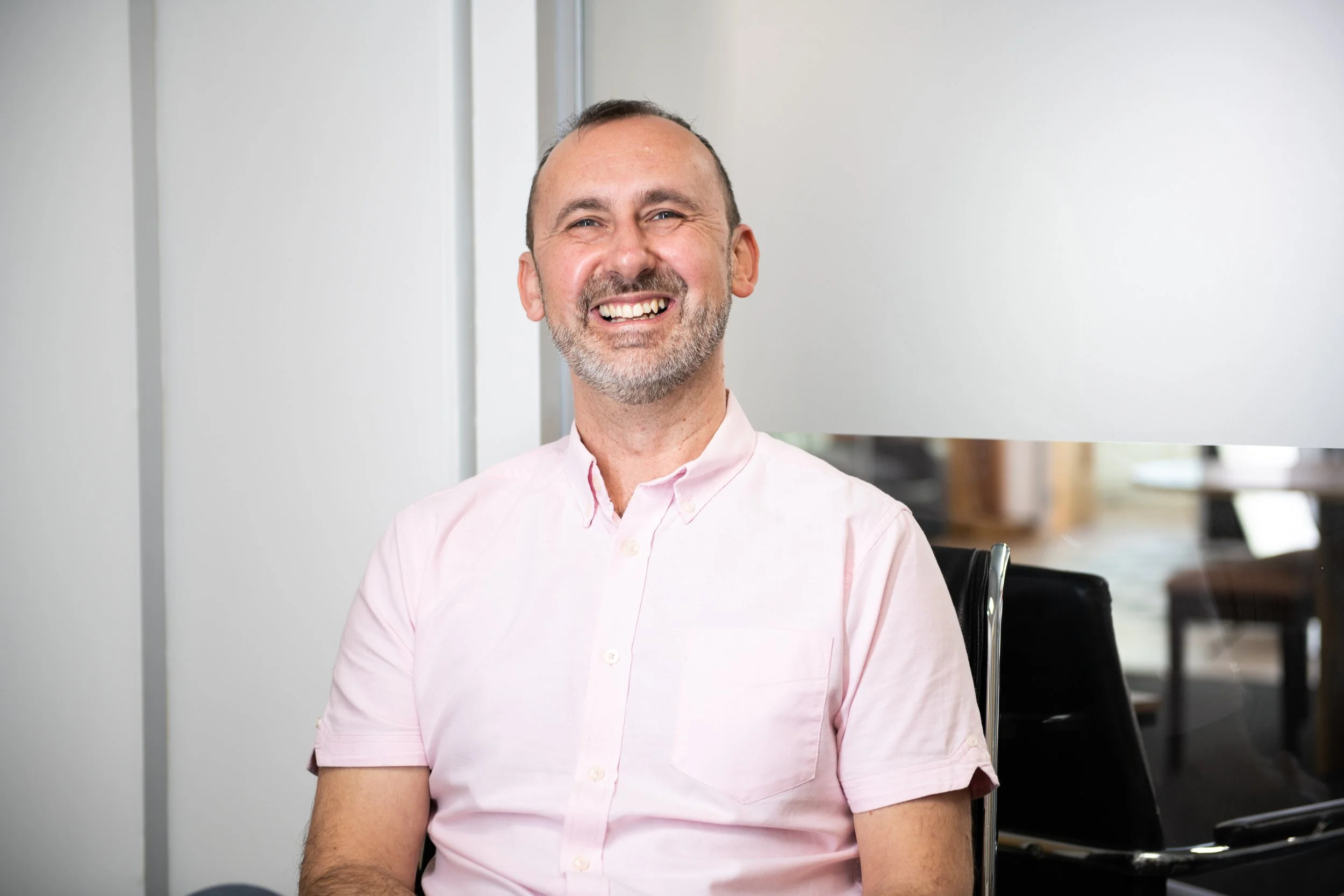 A smiling man with a closely trimmed beard and a receding hairline, wearing a light pink button-up shirt, sitting in a modern room with white walls and some black chairs.