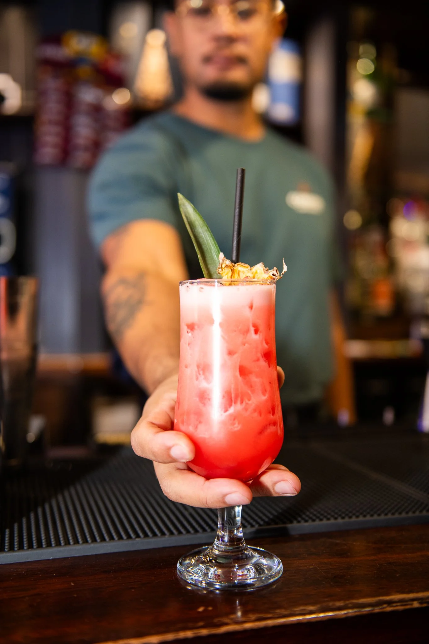 A person is holding a pinkish-red tropical cocktail with a pineapple leaf and fruit garnish, in a tall glass, with a black straw, in a bar setting.