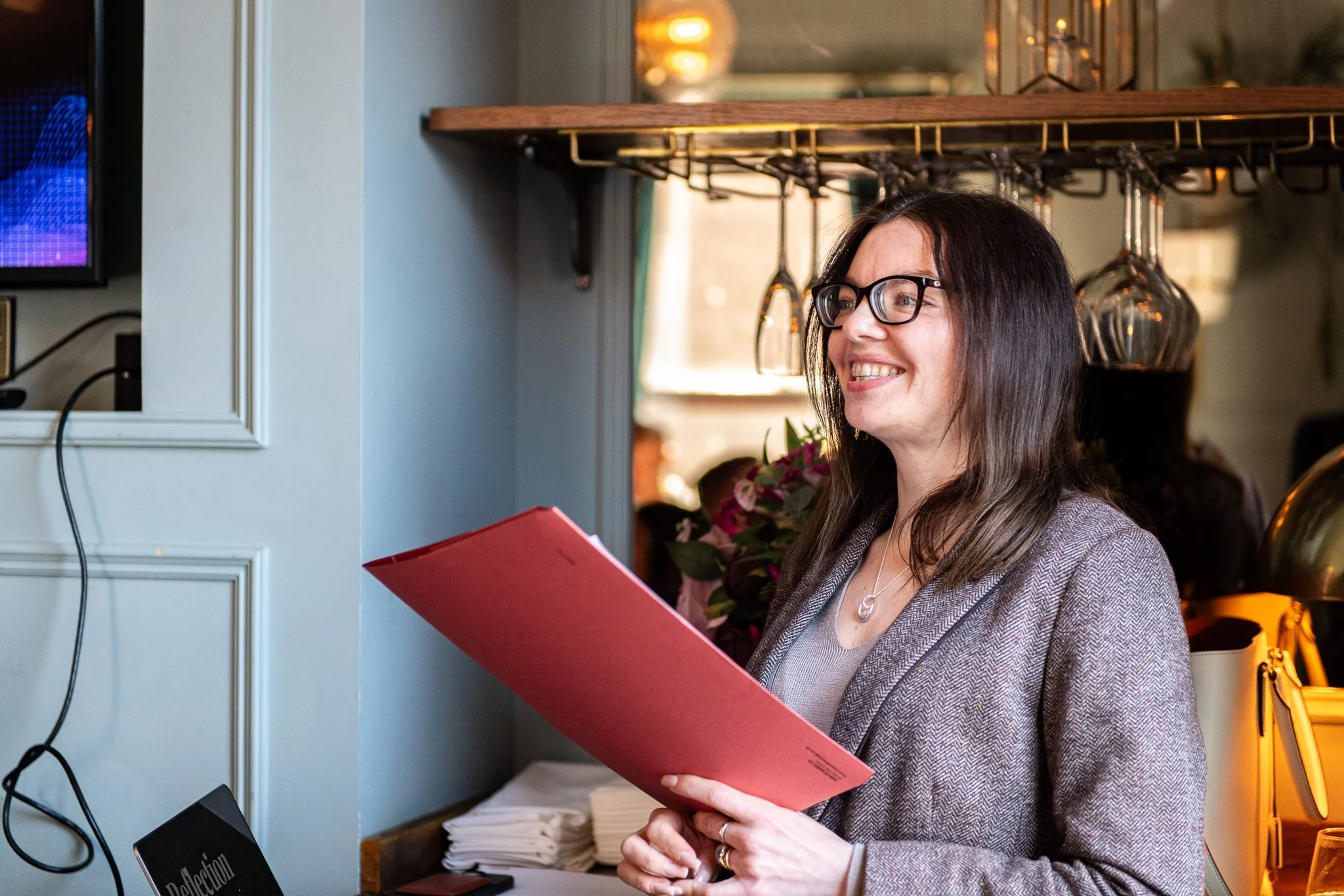 A smiling woman with long brown hair, glasses, and a checkered blazer holding a red menu inside a restaurant or cafe. There are hanging wine glasses and a bouquet of flowers behind her, and a low table with neatly stacked white napkins.