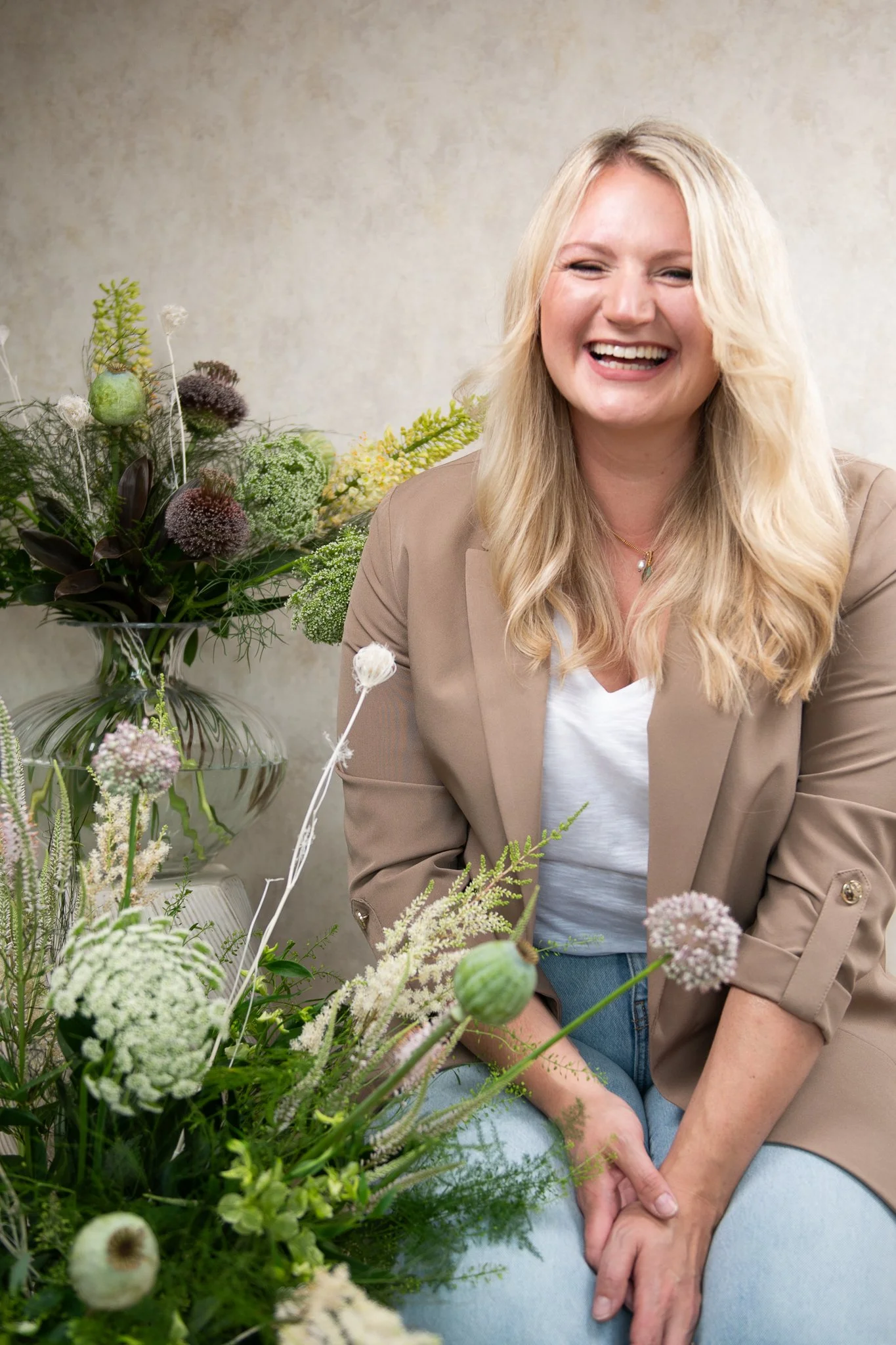 A woman with long blonde hair smiling while sitting among a variety of flowers and greenery.