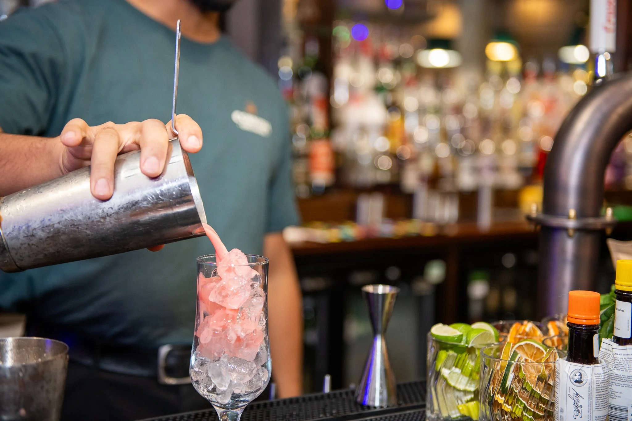 Bartender pouring pink cocktail over ice into a tall glass at a bar, with various bottles and bar tools in the background.
