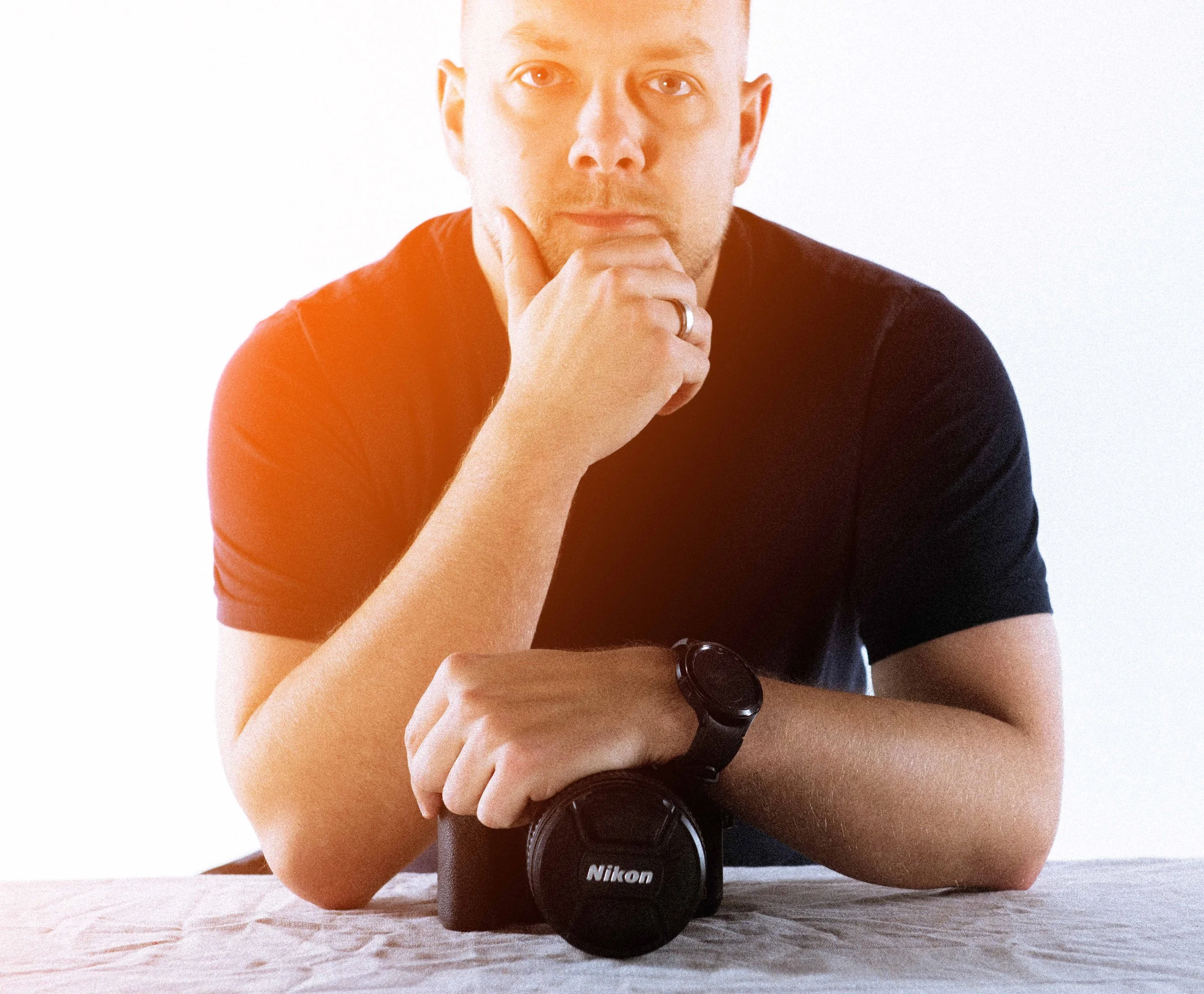 A person in a black shirt with a watch on their wrist is resting their chin on their hand, looking directly at the camera. A Nikon camera is placed on the table in front of them.