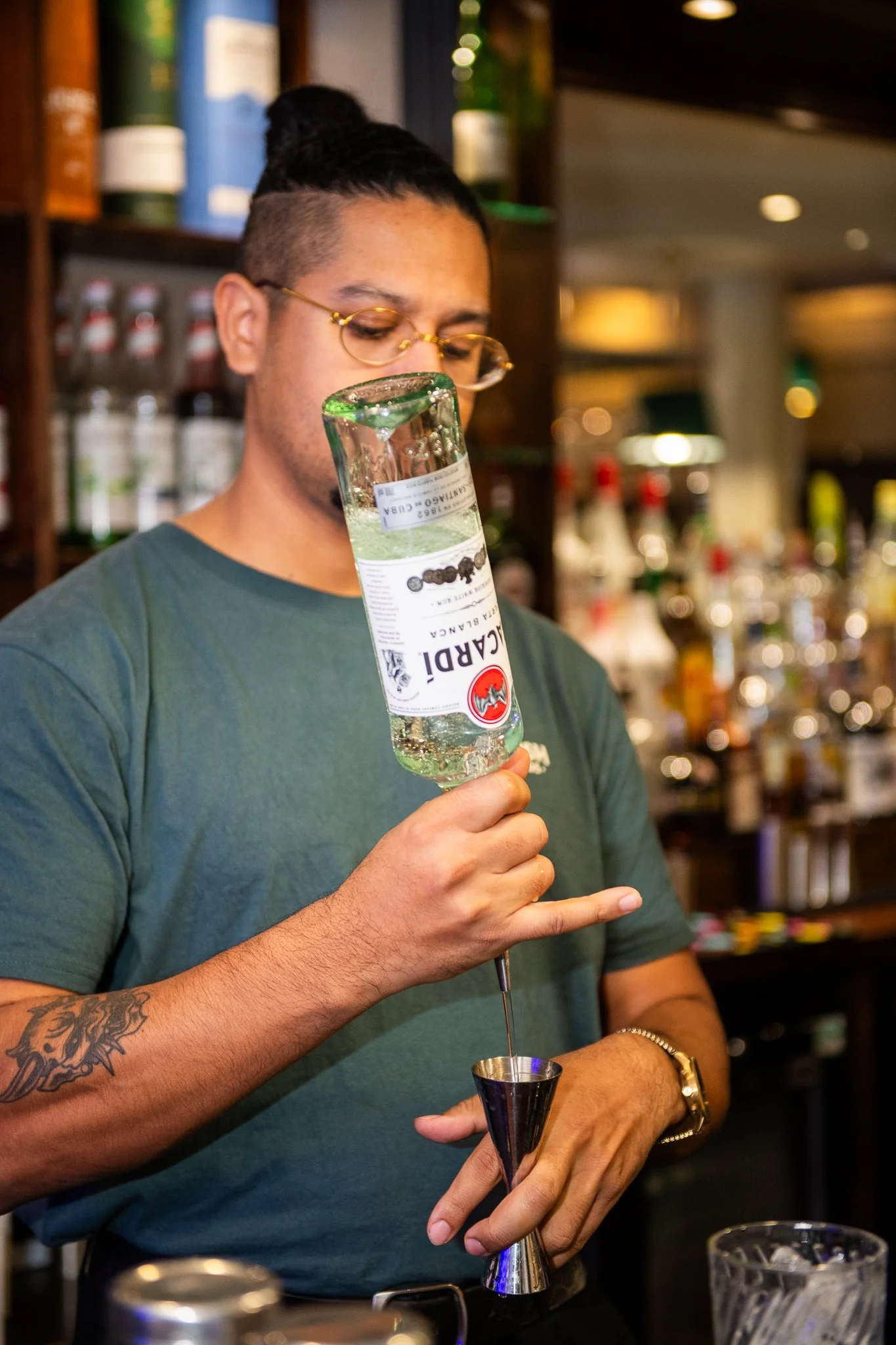 A person with glasses and a bun hairstyle is pouring clear liquid from a bottle into a jigger at a bar. They have a tattoo on their arm and are wearing a bracelet and watch. Shelves with bottles are visible in the background.