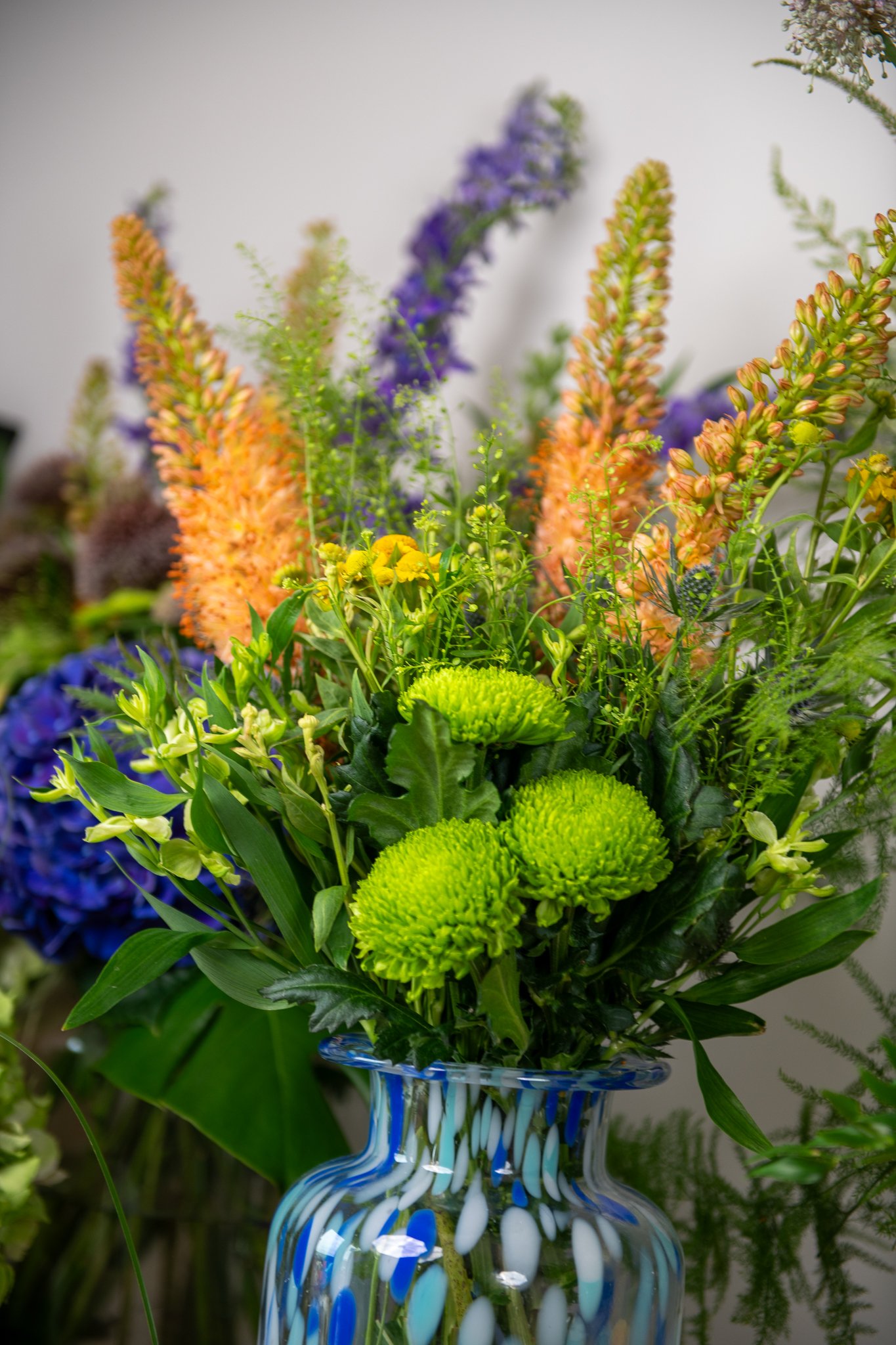 A bouquet of colorful flowers in a blue and white glass vase, including green, purple, yellow, and orange flowers with lush green foliage.