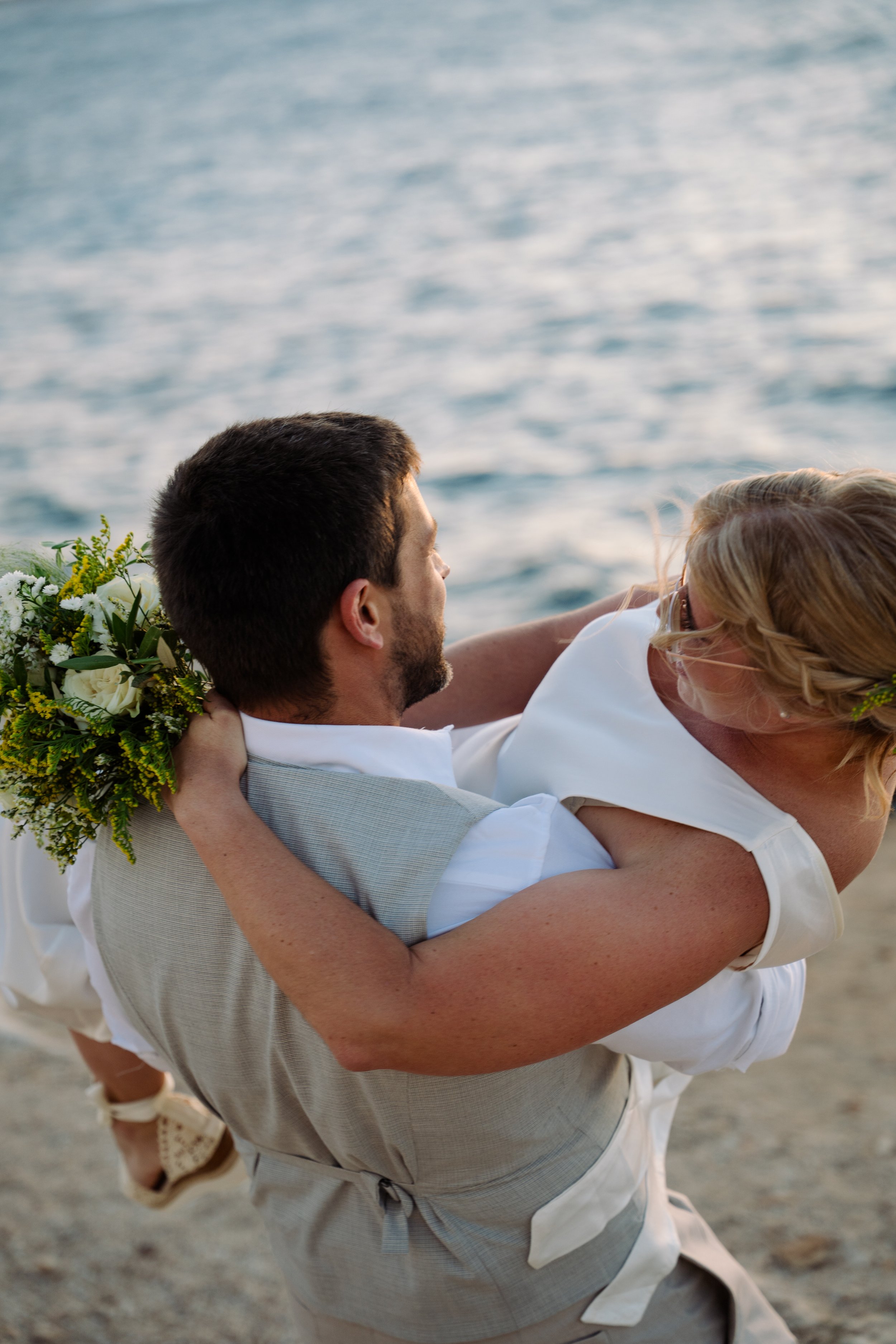 A man wearing a beige vest and white shirt is holding a woman with blonde hair in a white dress, lifting her in his arms near the water on a beach, with a bouquet of flowers visible in the background.