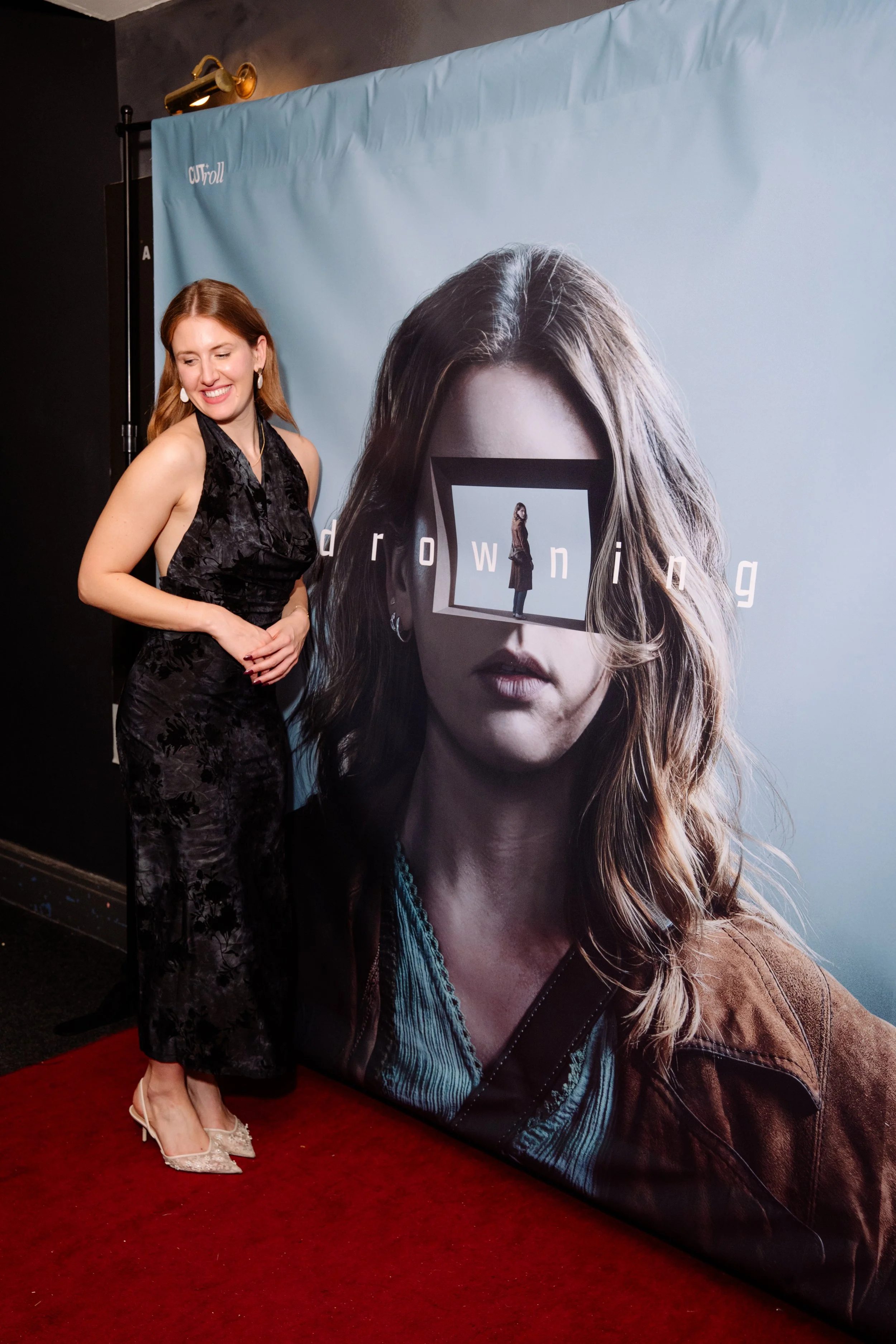 A woman in a black dress with floral pattern and white high heels standing on a red carpet next to a promotional poster for the show 'Drowning'.