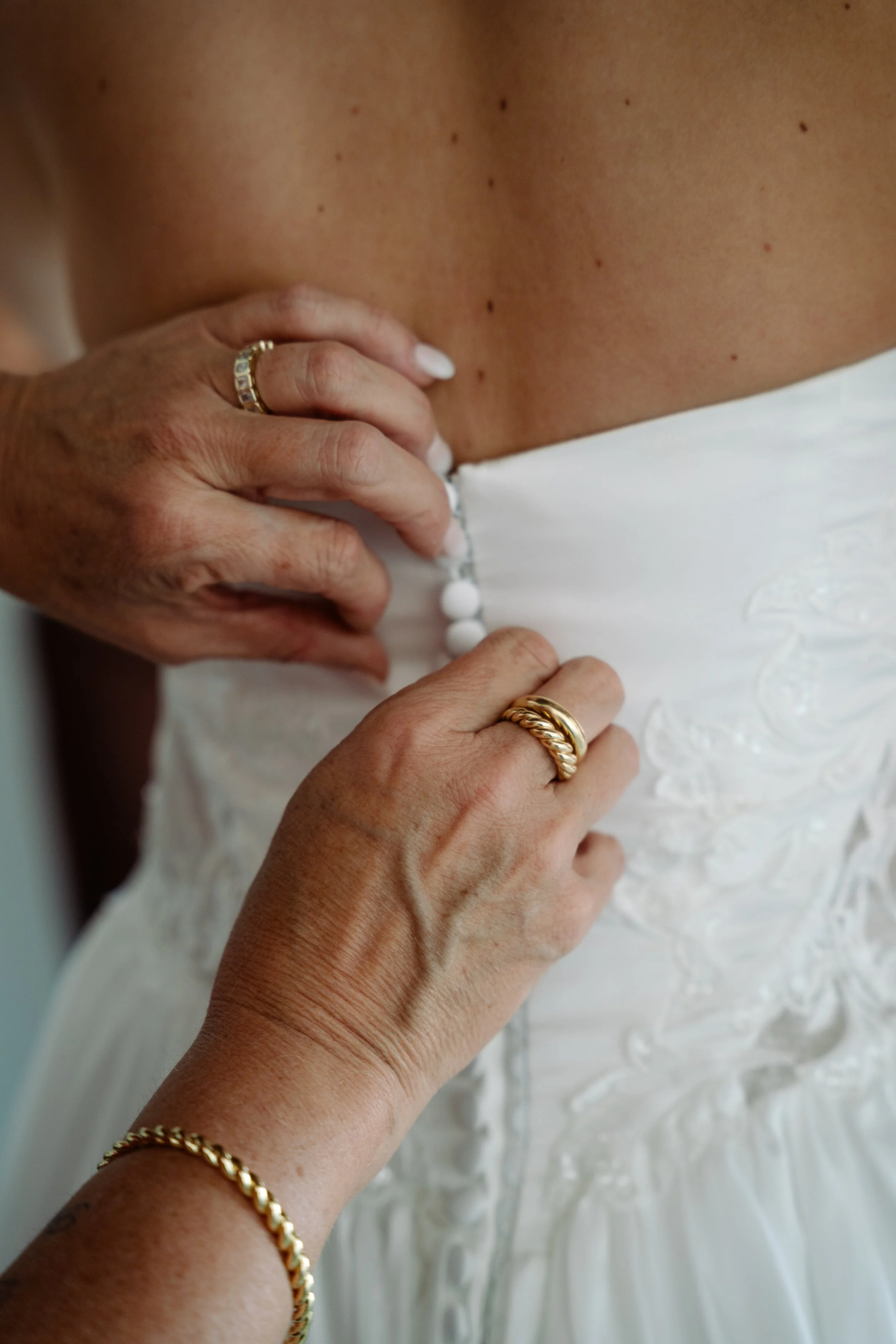 Close-up of a woman's back and hand, showing a white wedding dress with buttons, wearing gold jewelry, and the woman has light skin with freckles.