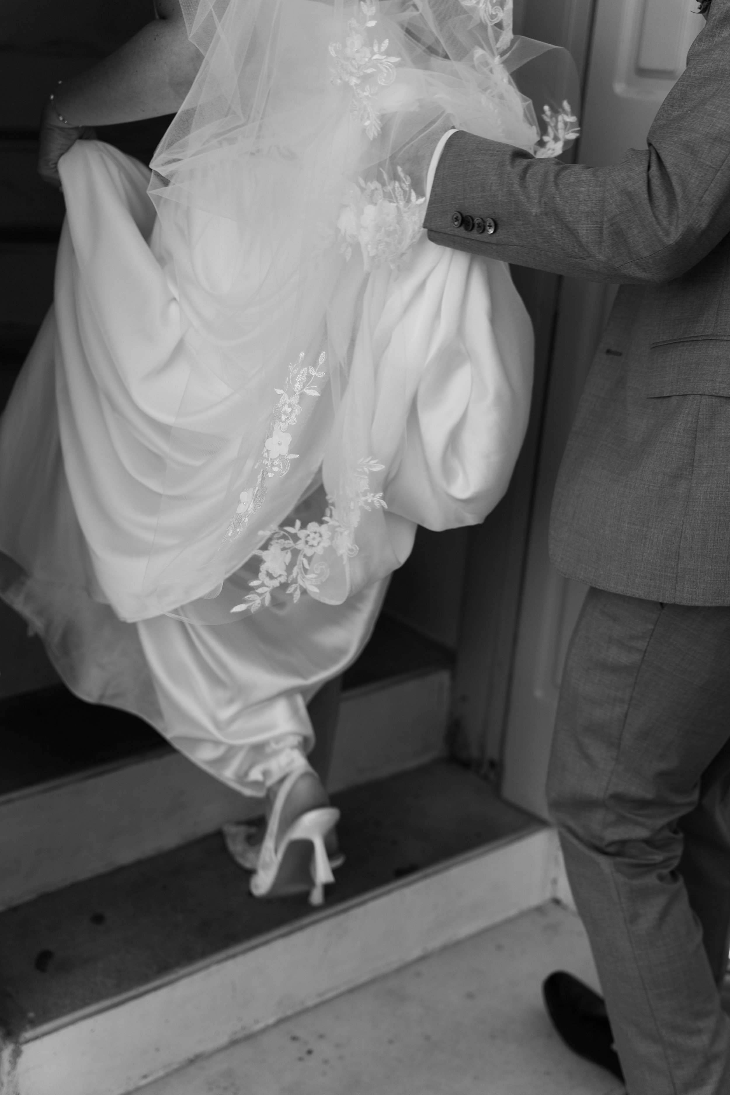 Black and white photo showing a bride in a wedding dress and veil, getting help putting on her shoe, with a man in a suit assisting her.