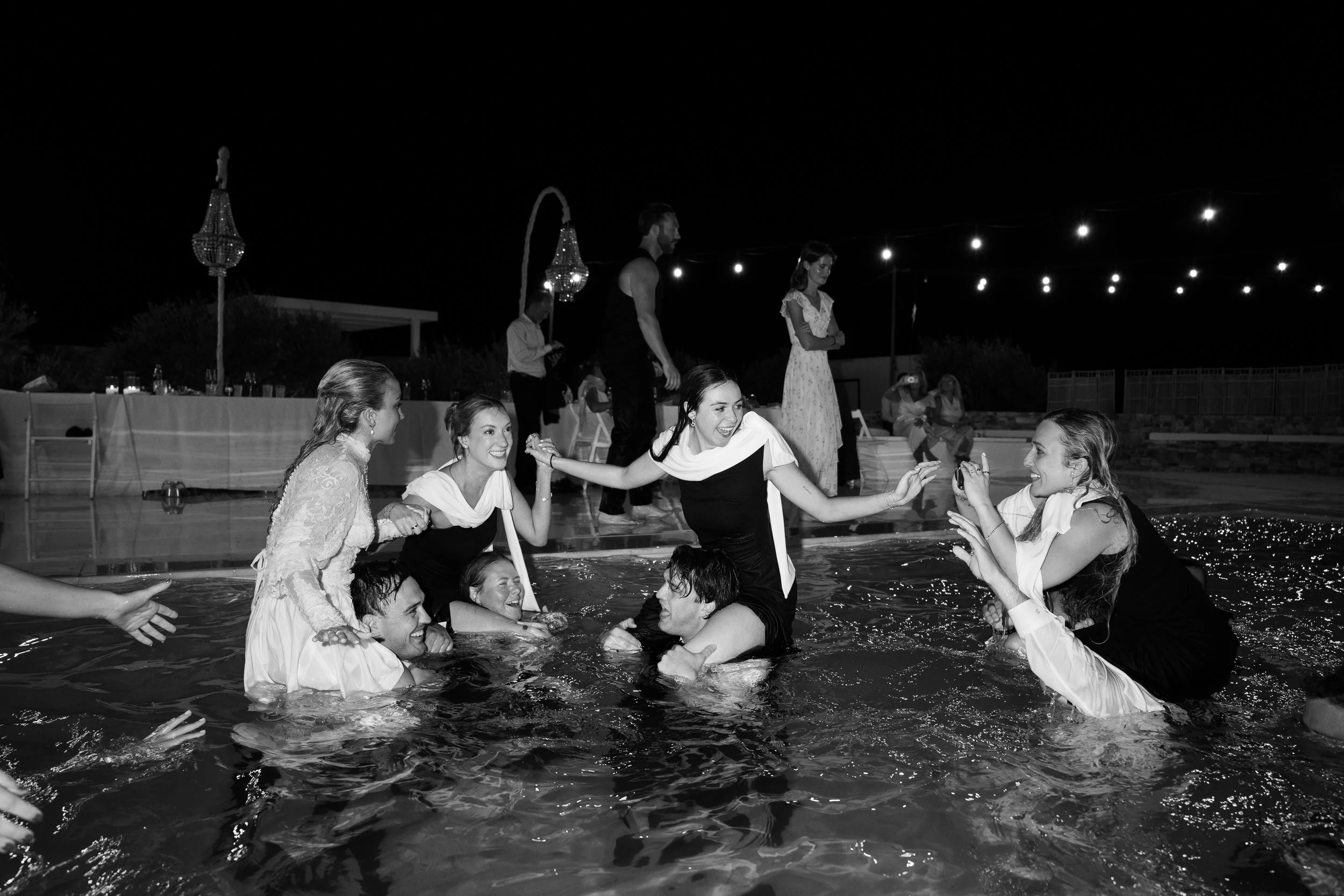 Group of women in evening dresses having fun in a swimming pool at night, with string lights and chandeliers overhead.