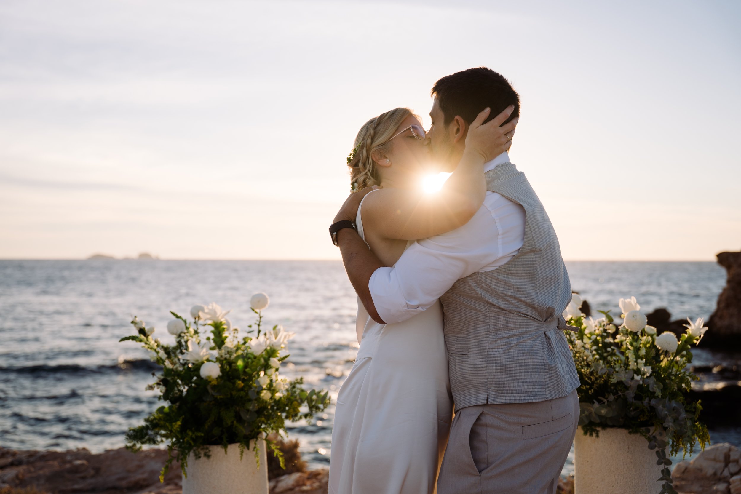 A couple sharing a kiss by the waterfront during sunset, with the woman wearing glasses and the man in a vest, surrounded by white flowers.