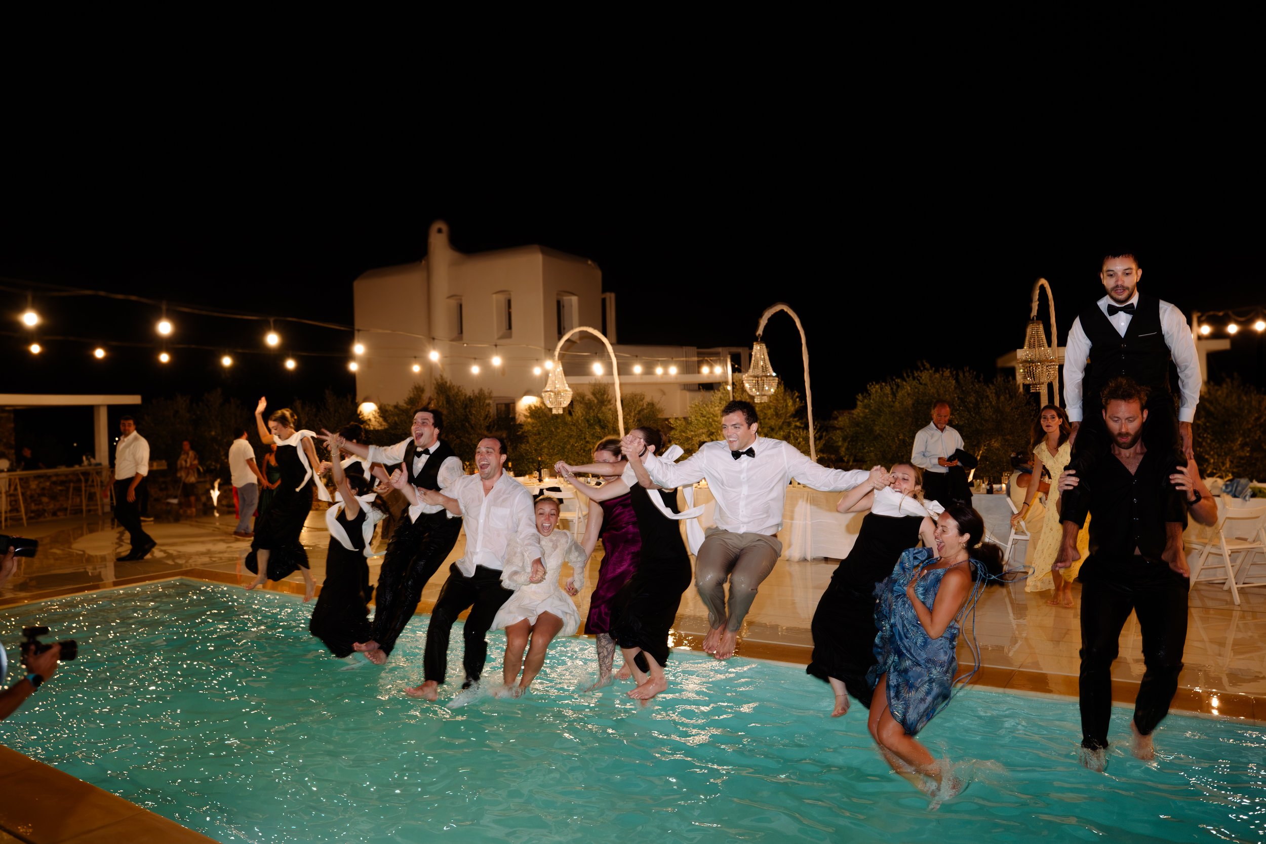 People in formal attire participating in a wedding reception, jumping into a swimming pool at night, with string lights and chandeliers overhead.