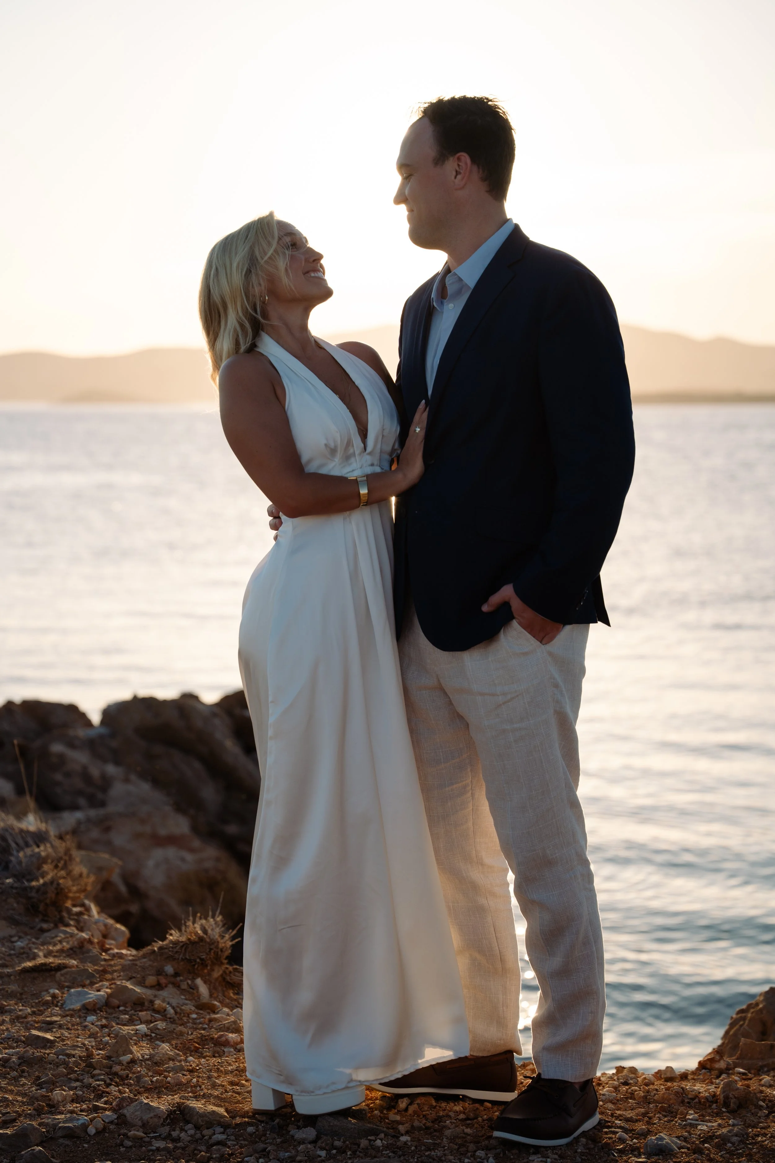 A couple dressed for a special occasion standing close together on a rocky shoreline during sunset, with a body of water and distant hills in the background.